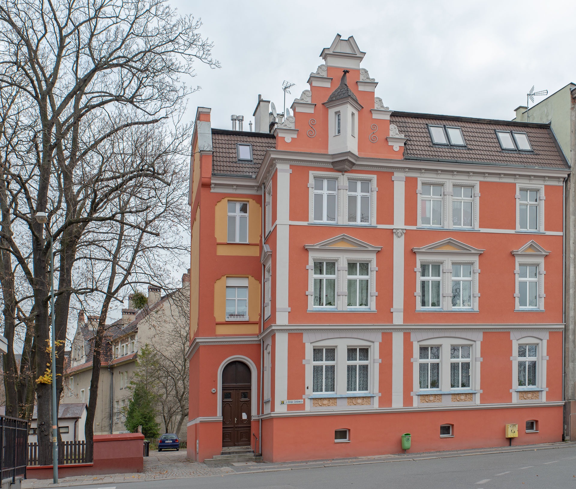 Odrestaurowana kamienica z pomarańczową elewacją, białymi zdobieniami okien i ozdobnym szczytem, widziana z perspektywy ulicy w pochmurny dzień.