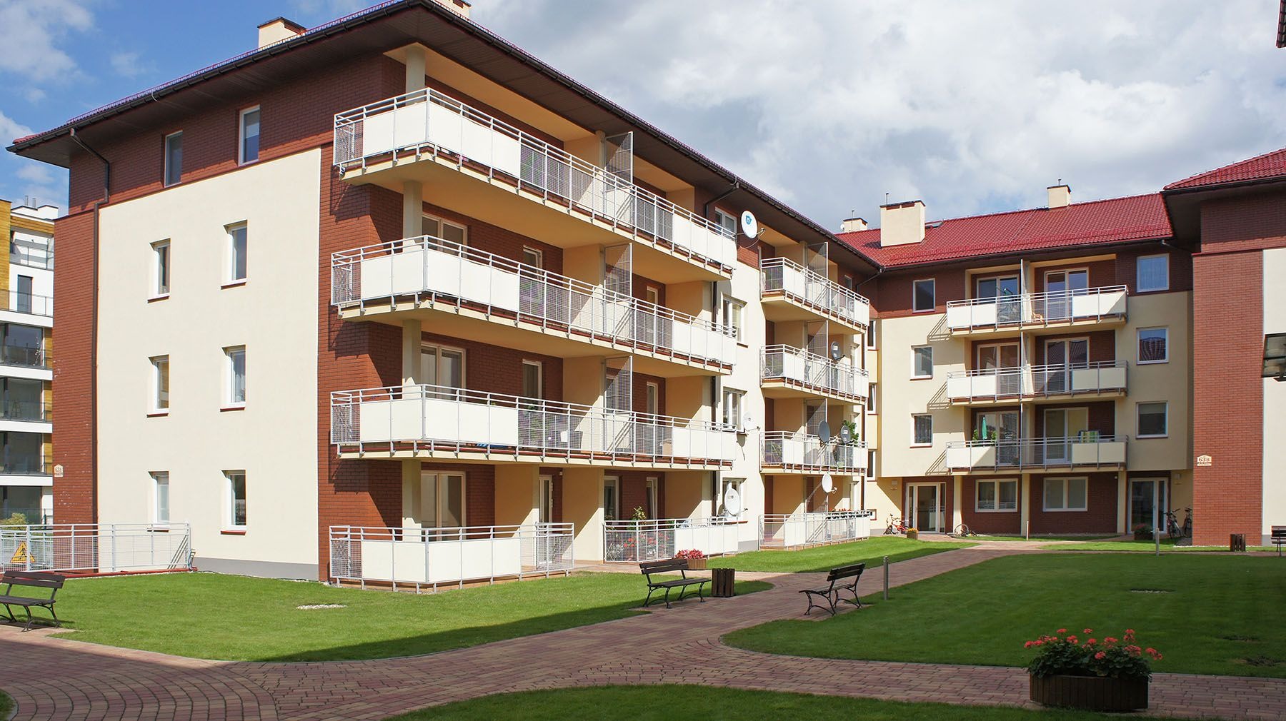 Nowoczesny budynek wielorodzinny z elewacją w odcieniach beżu i czerwonej cegły, z balkonami z białymi balustradami, widok z wewnętrznego dziedzińca z ławkami i zadbanym trawnikiem.