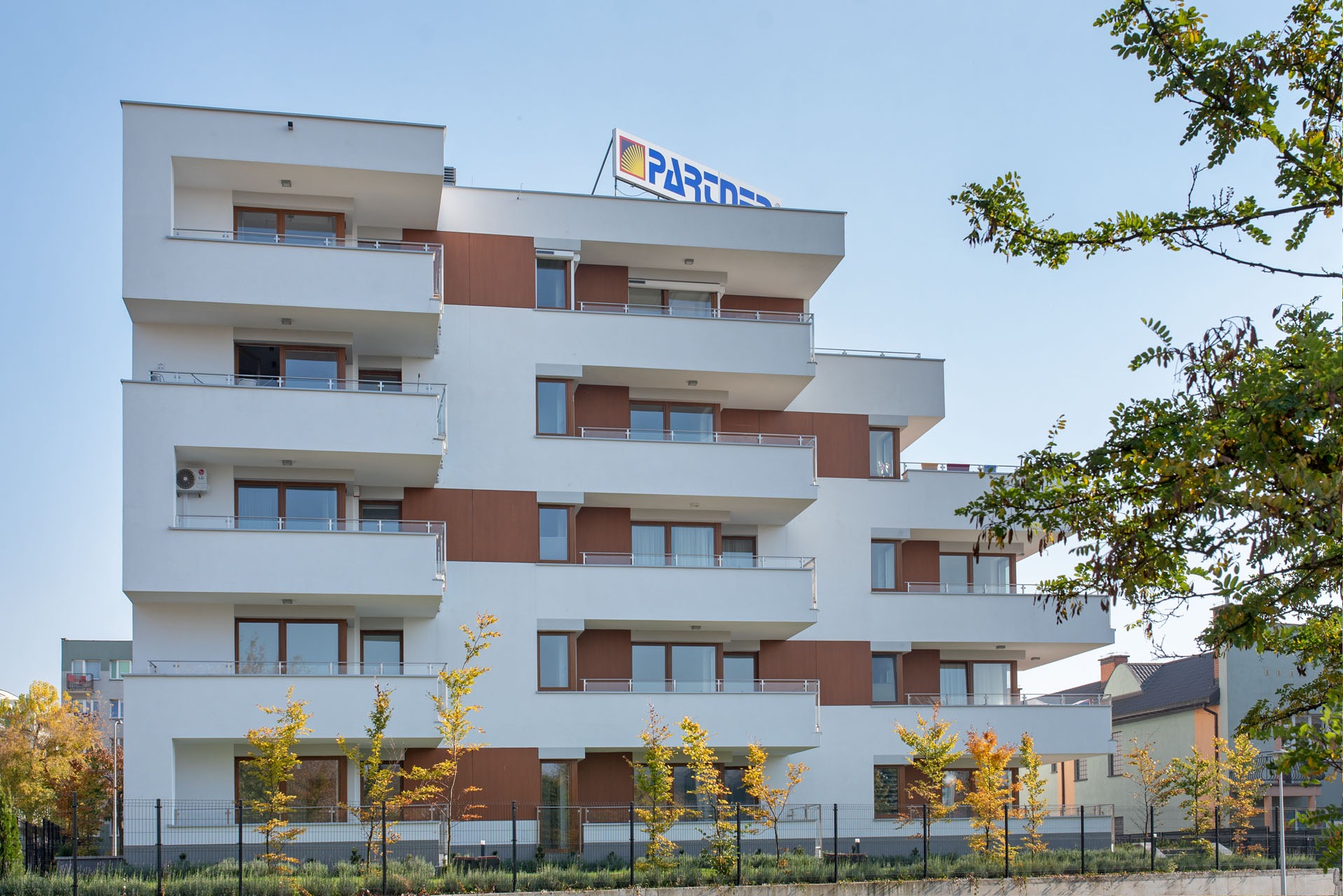 Nowoczesny budynek wielorodzinny z białymi ścianami, brązowymi panelami i balkonami, z logo firmy na dachu, otoczony zielenią.