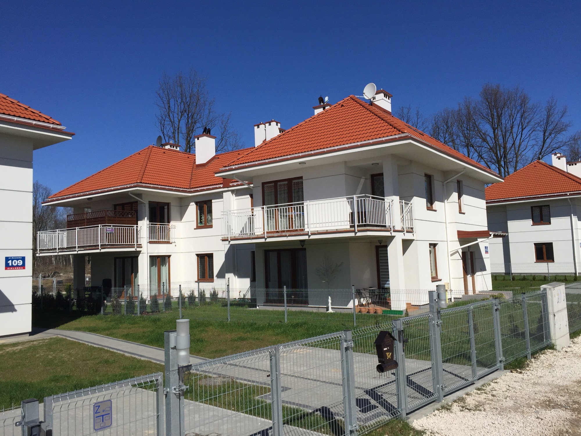 Nowoczesne, białe budynki z czerwonymi dachami krytymi dachówką, ogrodzone metalowym płotem, z numerem domu 109 na elewacji, widoczne w słoneczny dzień.