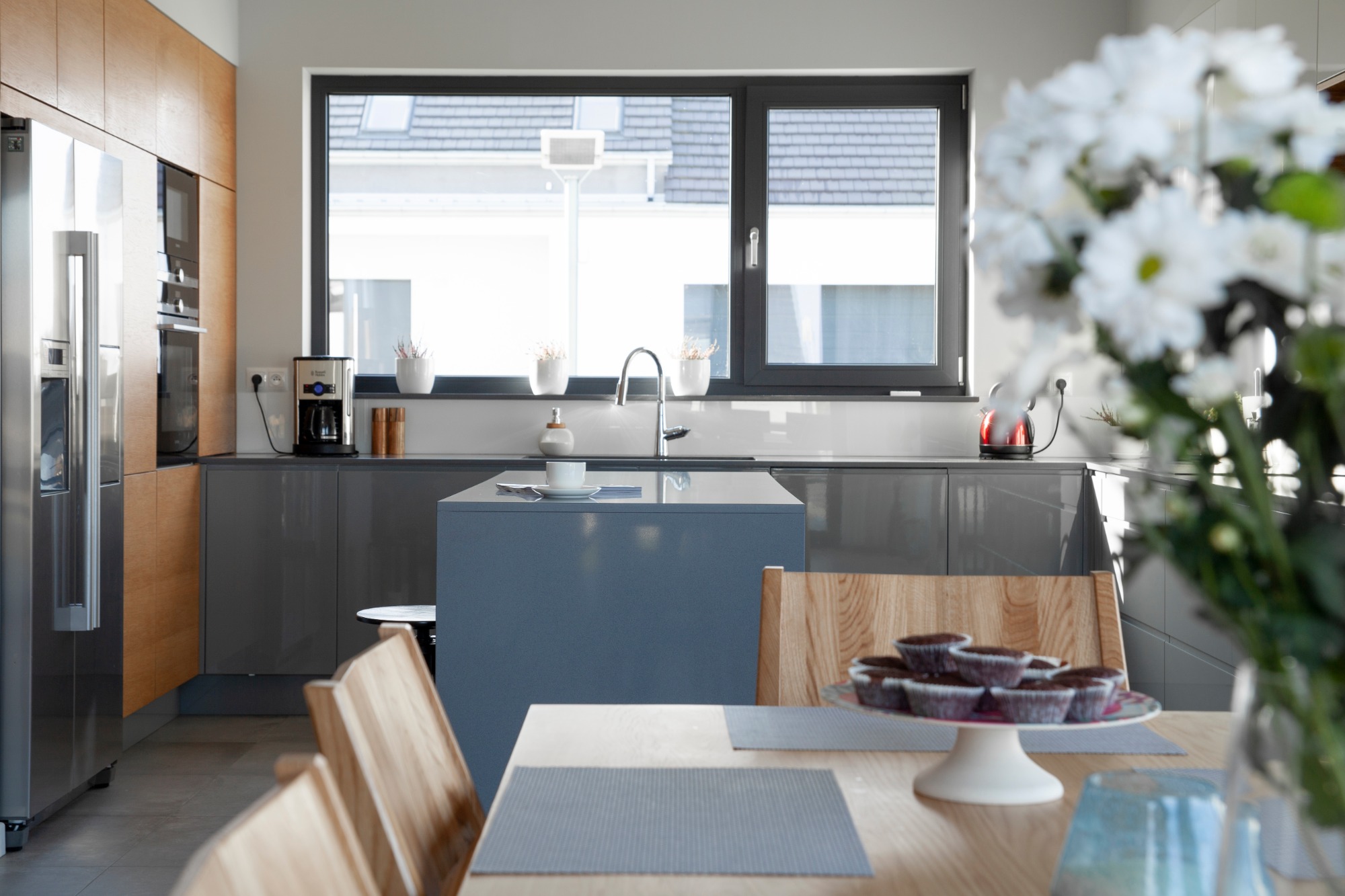 Nowoczesna kuchnia z szarymi lakierowanymi szafkami, drewnianymi akcentami i wyspą. Na stole talerz z babeczkami i wazon z białymi kwiatami.