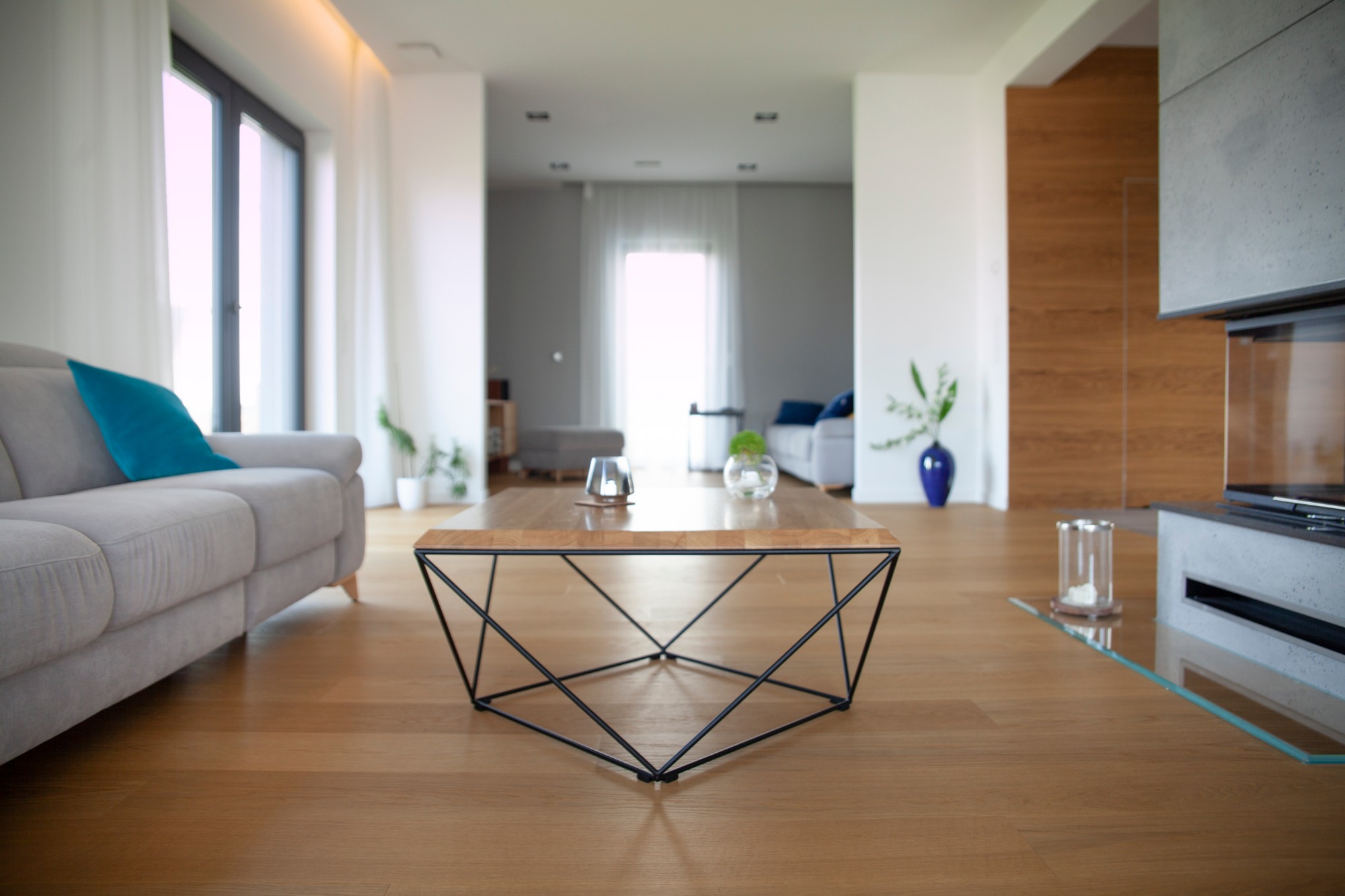 Jasny salon z drewnianą podłogą, szarą sofą z turkusową poduszką i geometrycznym stolikiem kawowym na pierwszym planie, w tle widoczny kominek i dalsza część salonu.