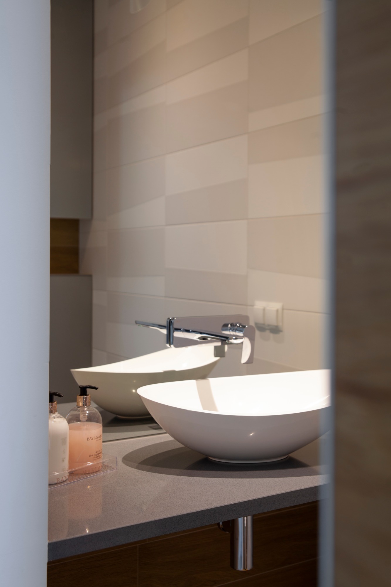 Nowoczesna łazienka z dwiema owalnymi umywalkami nablatowymi, chromowanymi bateriami, beżowymi płytkami na ścianie i minimalistycznymi dozownikami mydła.
