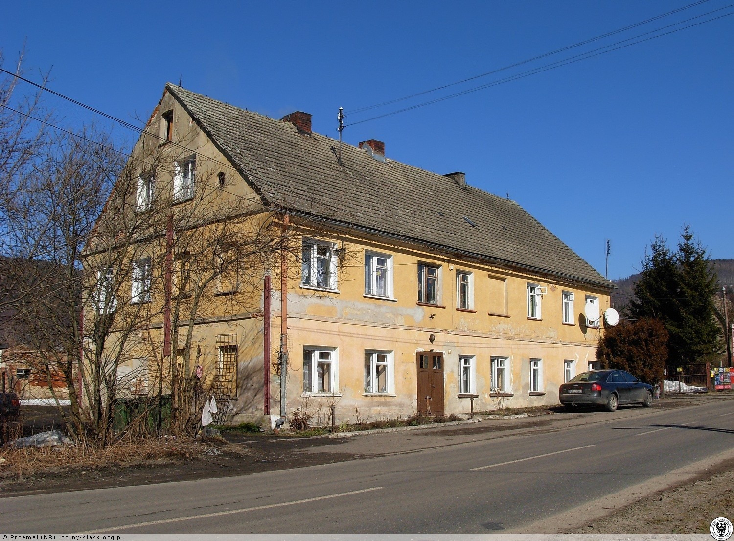 Dwukondygnacyjny budynek z żółtą, częściowo zniszczoną elewacją, widoczny z ulicy w słoneczny dzień. Na elewacji widoczne ślady wcześniejszych prac, w tle góra i drzewa.
