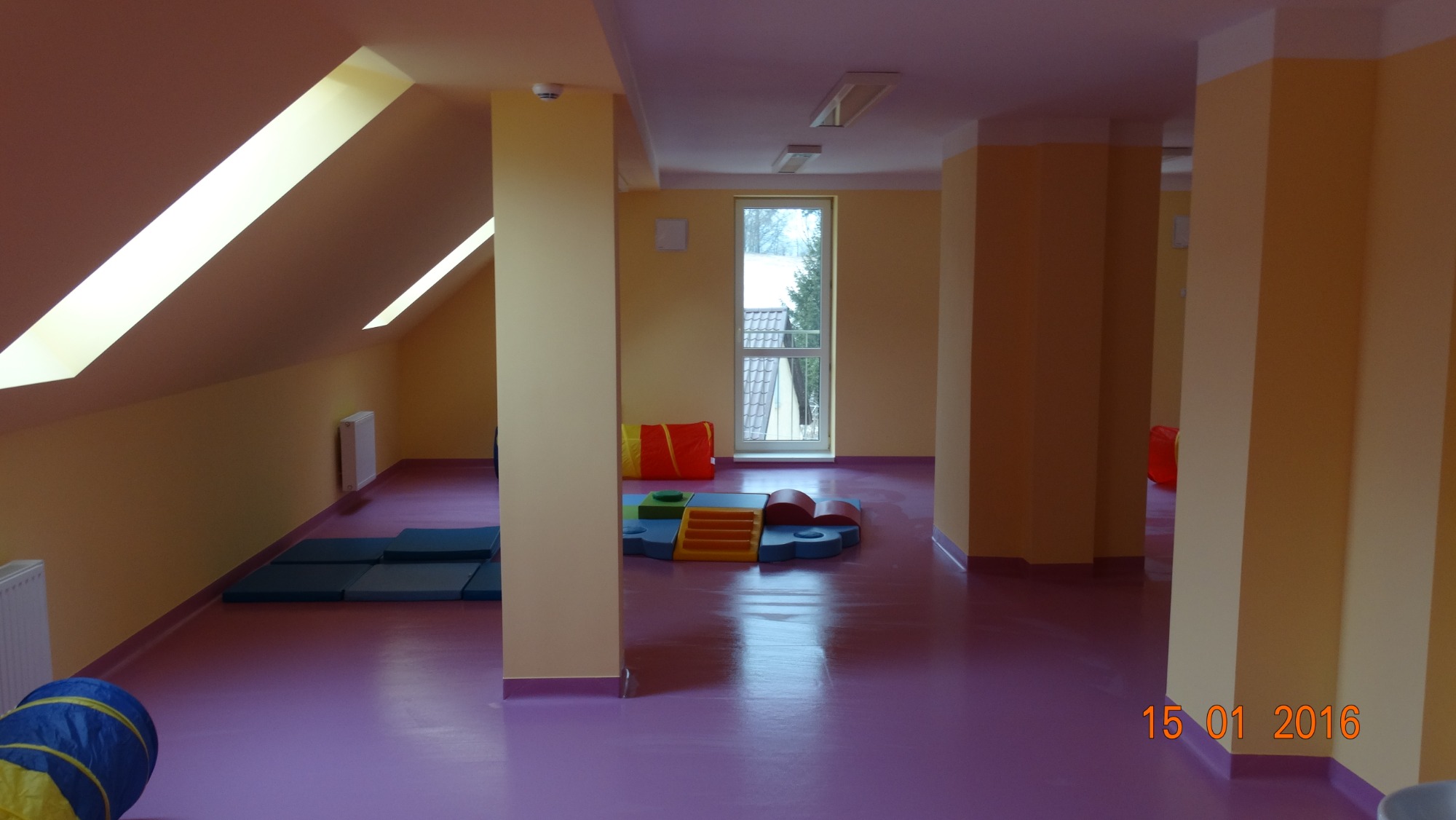 Jasne wnętrze sali zabaw na poddaszu z fioletową podłogą, żółtymi ścianami i kolumnami, z oknem w tle i kolorowymi elementami wyposażenia.
