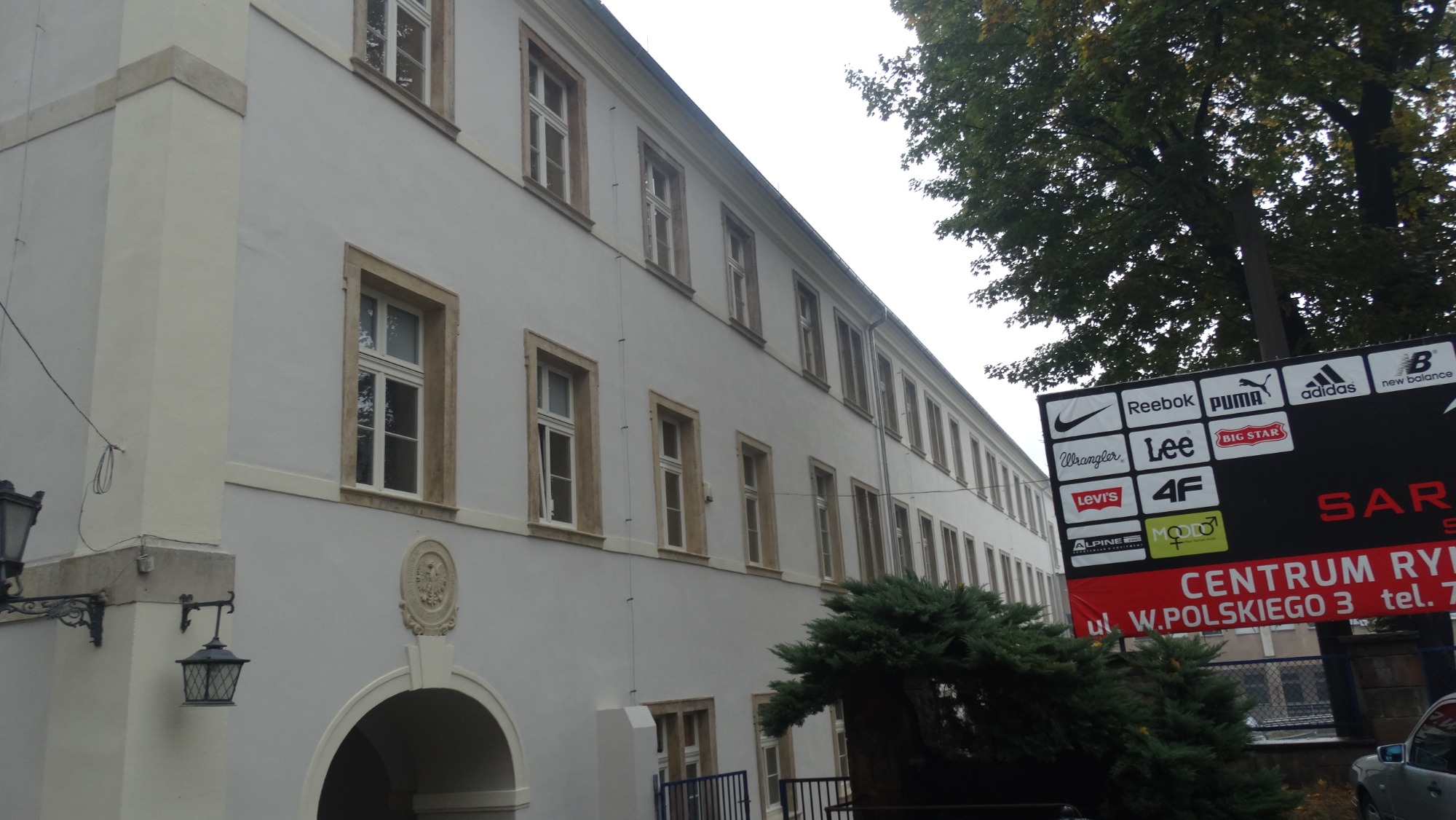 Długi budynek z jasną elewacją i oknami w kamiennych obramowaniach, widoczny z boku pod kątem, obok reklama z logotypami marek sportowych.
