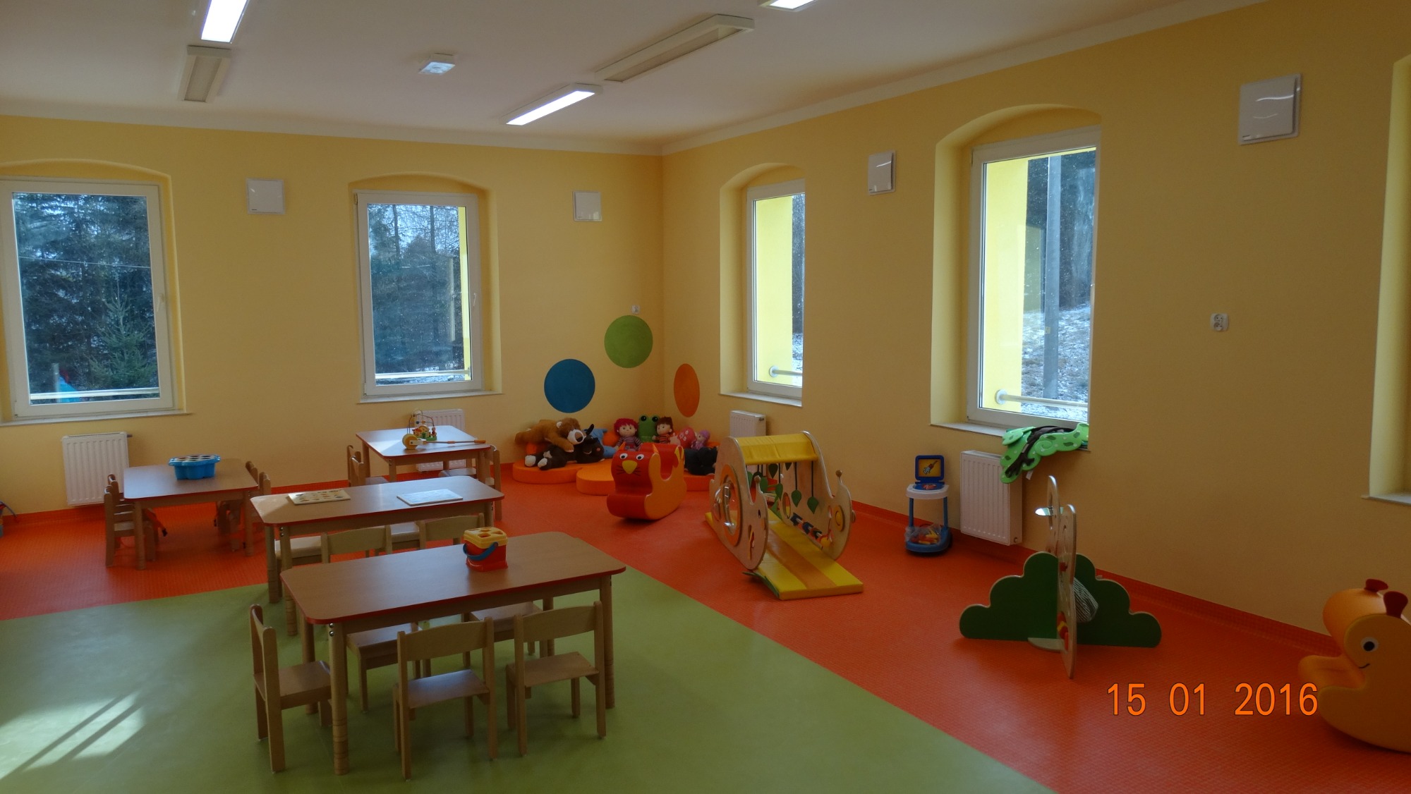 Jasne, nowo wykończone wnętrze sali przedszkolnej z żółtymi ścianami, drewnianymi stolikami i krzesłami, oraz kolorową podłogą z elementami pomarańczowymi i zielonymi, widoczne zabawki i dekoracje...