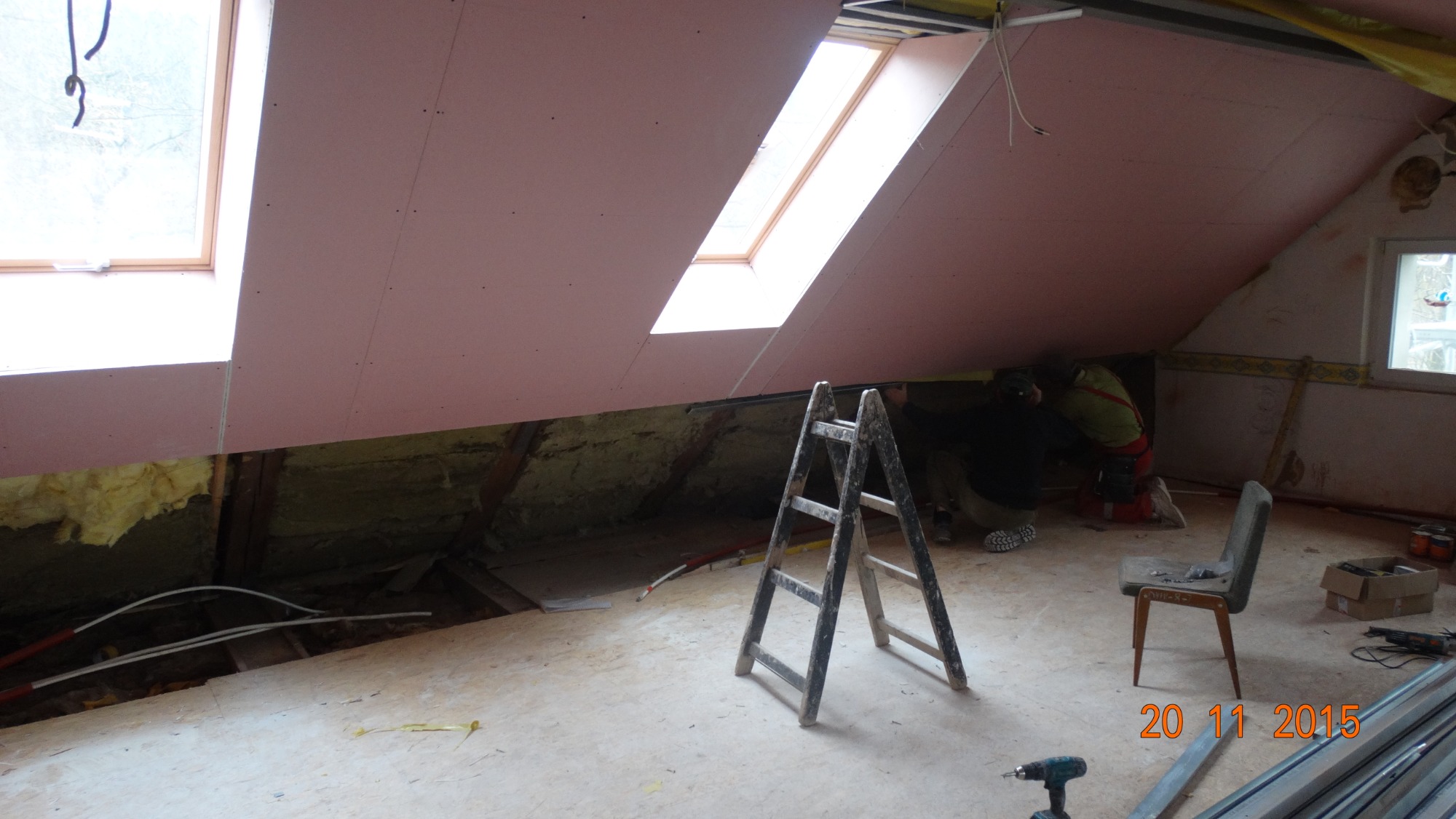 Montaż różowej płyty karton-gips na poddaszu ze skosami i oknami dachowymi, widoczna drabina, narzędzia i częściowo ocieplenie, dwóch pracowników w trakcie pracy.