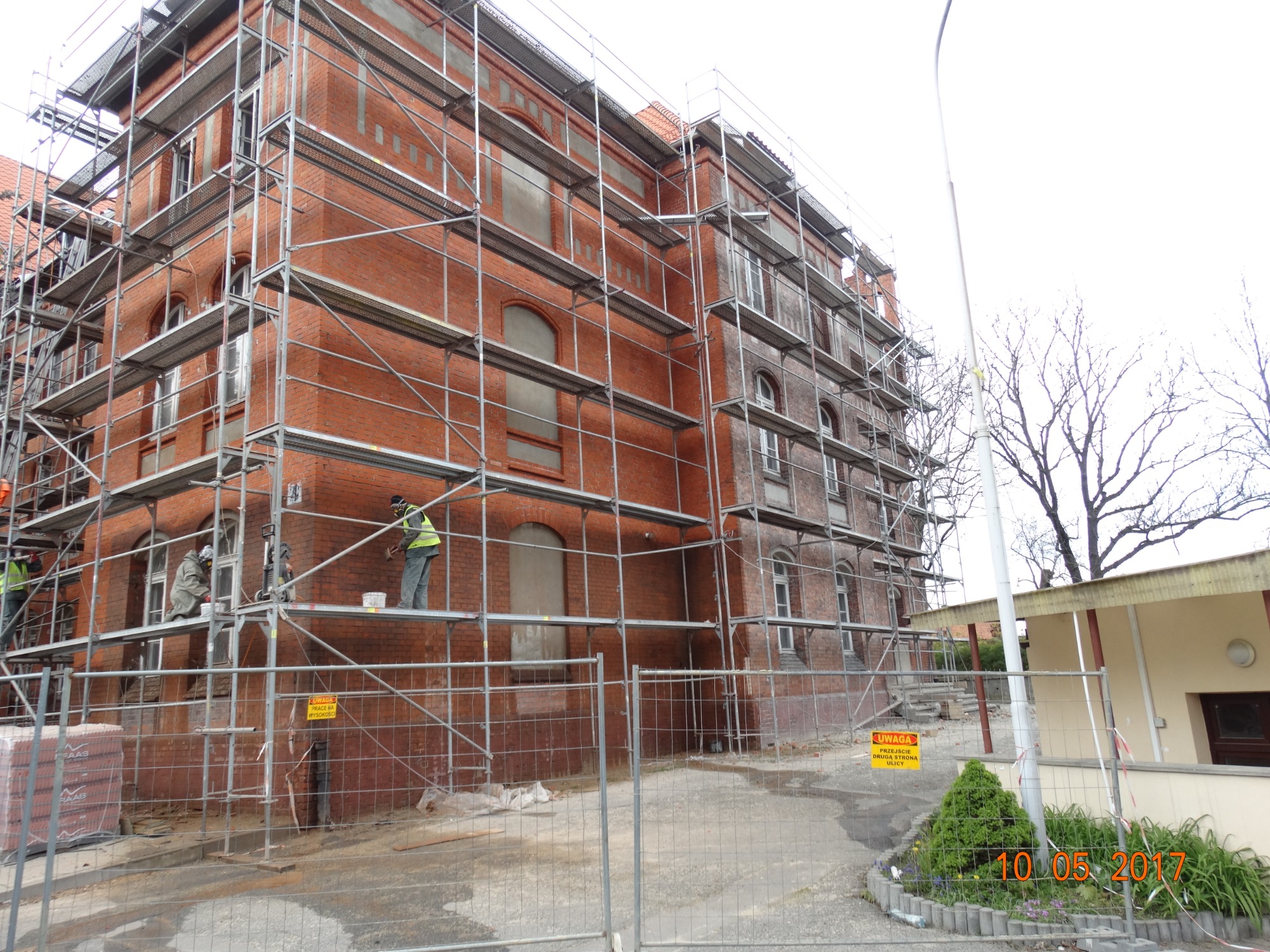 Renowacja ceglanej elewacji budynku z wykorzystaniem rusztowań, widoczni pracownicy w odblaskowych kamizelkach, częściowo otynkowane okna, paleta cegieł w tle, ogrodzenie placu budowy.