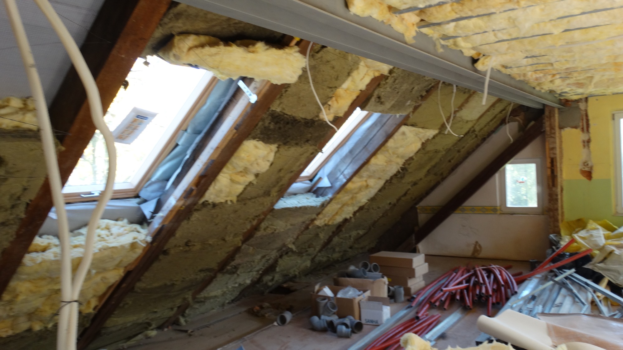 Poddasze w trakcie ocieplania, widoczne wełna mineralna między krokwiami, okno dachowe i elementy instalacji.