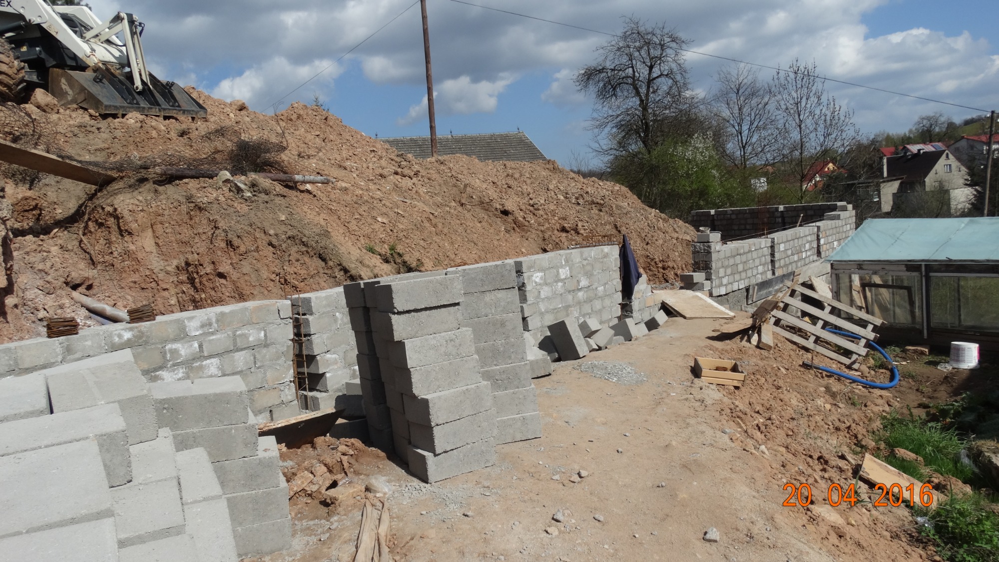 Budowa fundamentów z bloczków betonowych na nierównym terenie, widoczny kopiec ziemi i częściowo wzniesiona ściana, w tle zabudowania mieszkalne i maszyna budowlana.