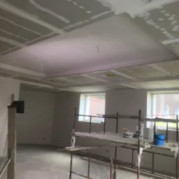 Montaż płyt kartonowo-gipsowych na suficie podwieszanym, widoczne rusztowanie i narzędzia w surowym wnętrzu z oknami.