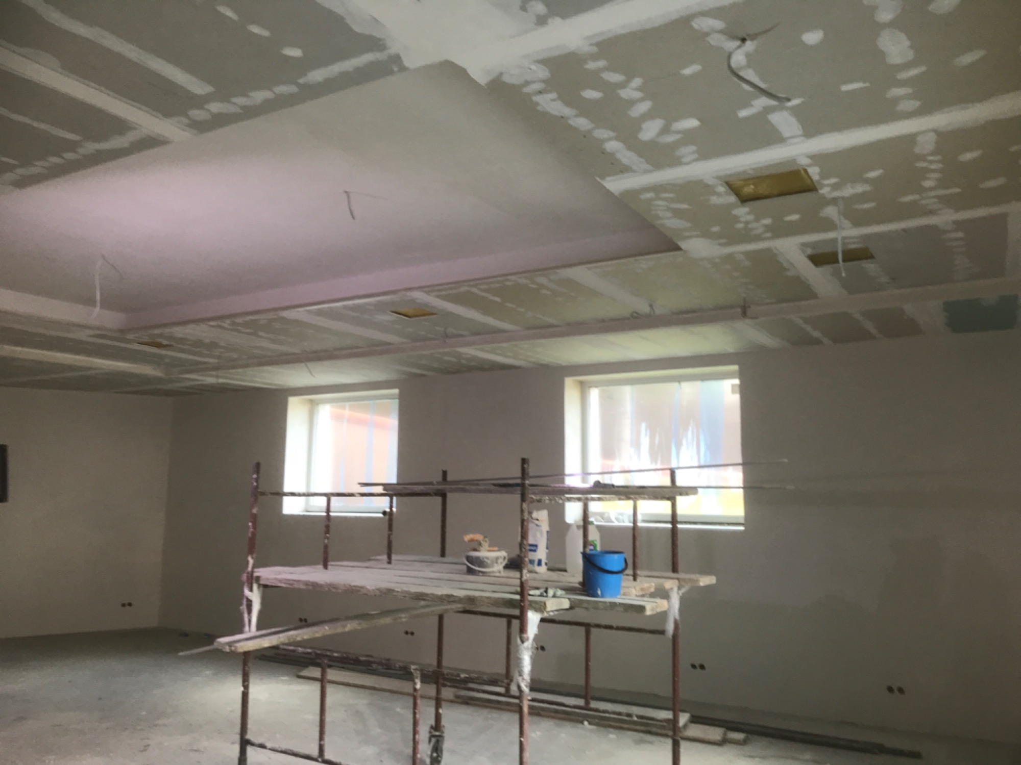 Wnętrze w trakcie wykańczania: sufit z płyt kartonowo-gipsowych, częściowo wyszpachlowany, widoczne rusztowanie z wiadrem i narzędziami, dwa okna w tle.