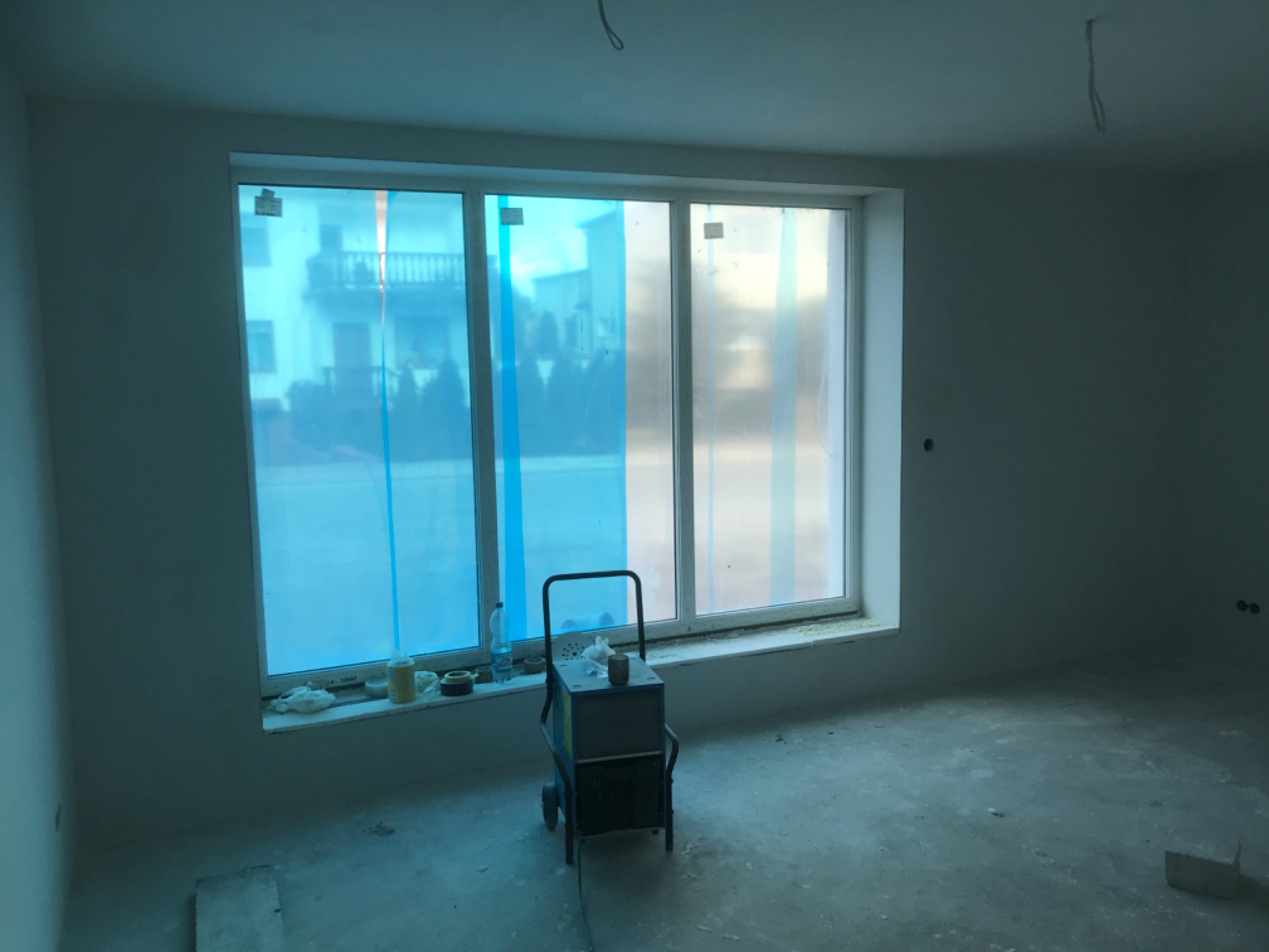 Wnętrze w trakcie remontu: okno zabezpieczone folią, na parapecie butelka wody i pojemniki, na podłodze maszyna na kółkach, surowy beton na podłodze.