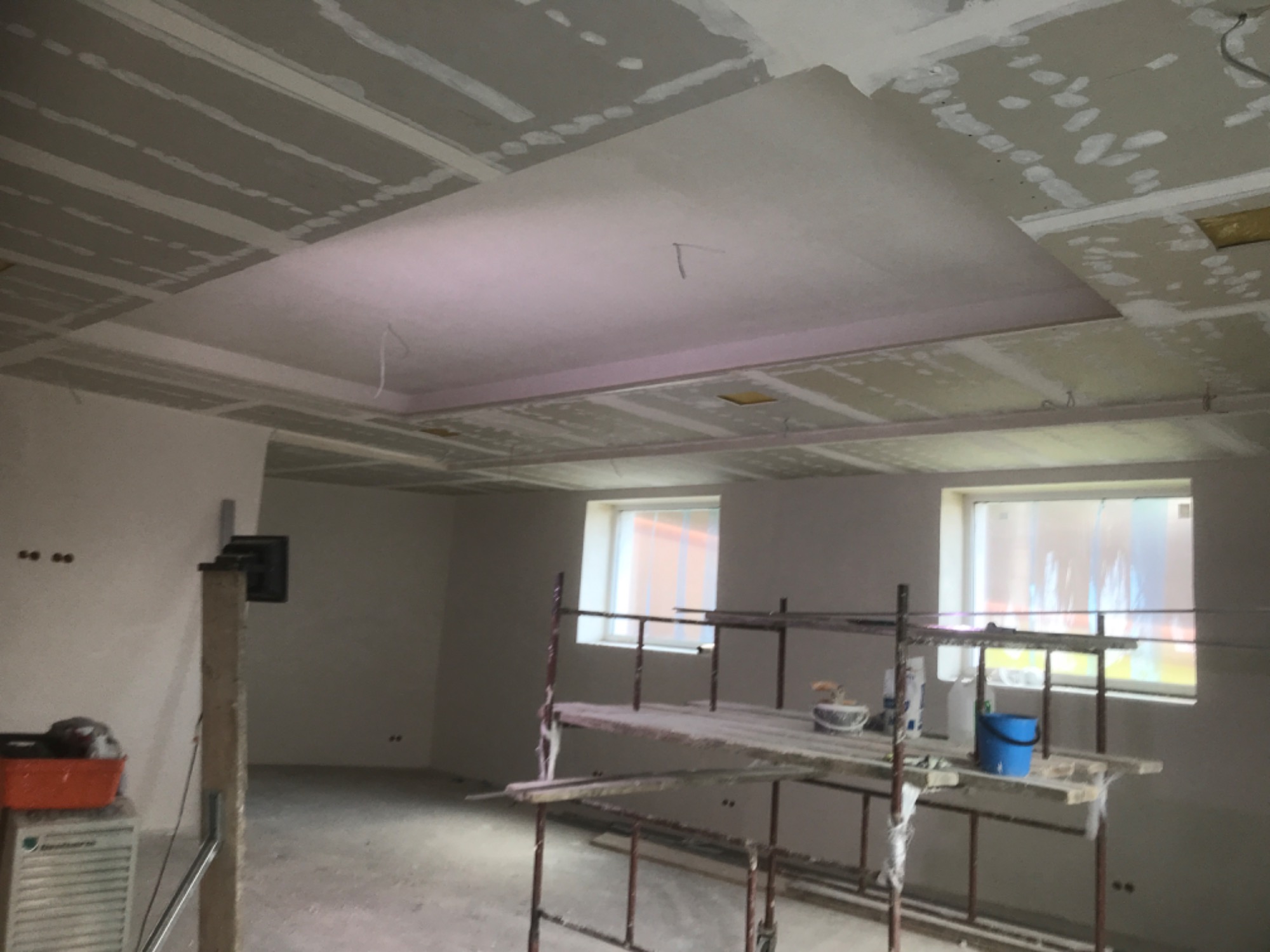 Montaż płyt kartonowo-gipsowych na suficie podwieszanym, widoczne rusztowanie i narzędzia w surowym wnętrzu z oknami.