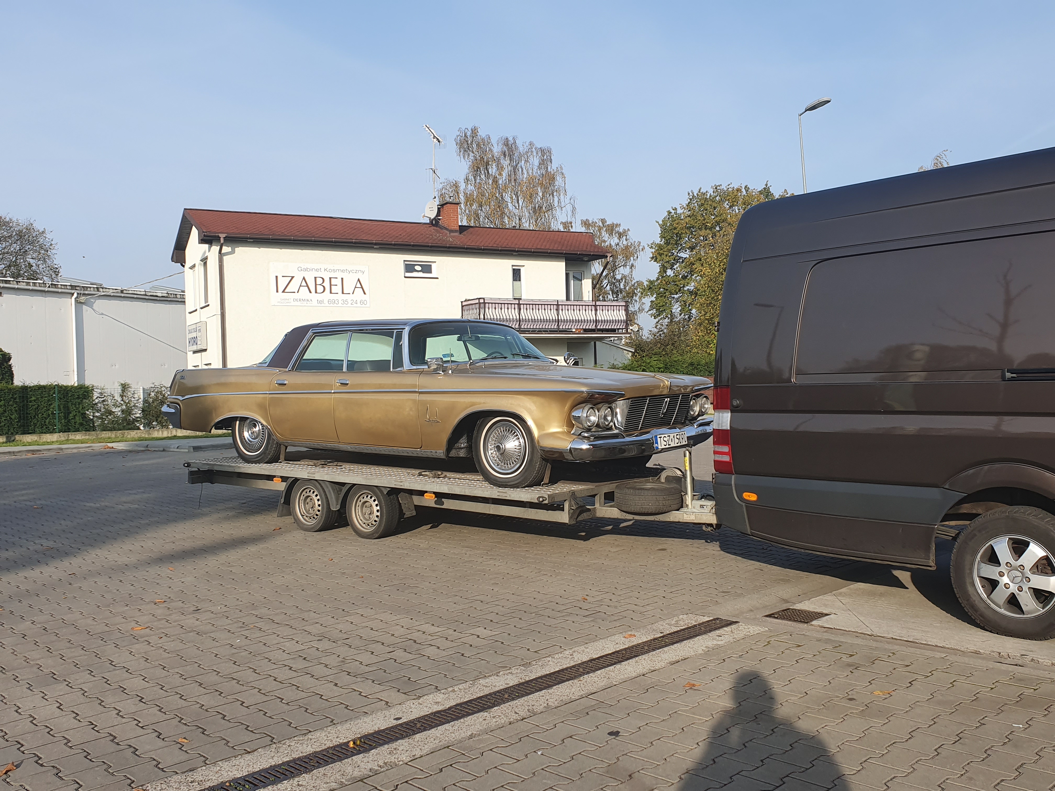 Złoty zabytkowy samochód marki Imperial z 1964 roku, przewożony na niskopodwoziowej przyczepie, podpiętej do brązowego busa na tle budynku z szyldem 'IZABELA'.