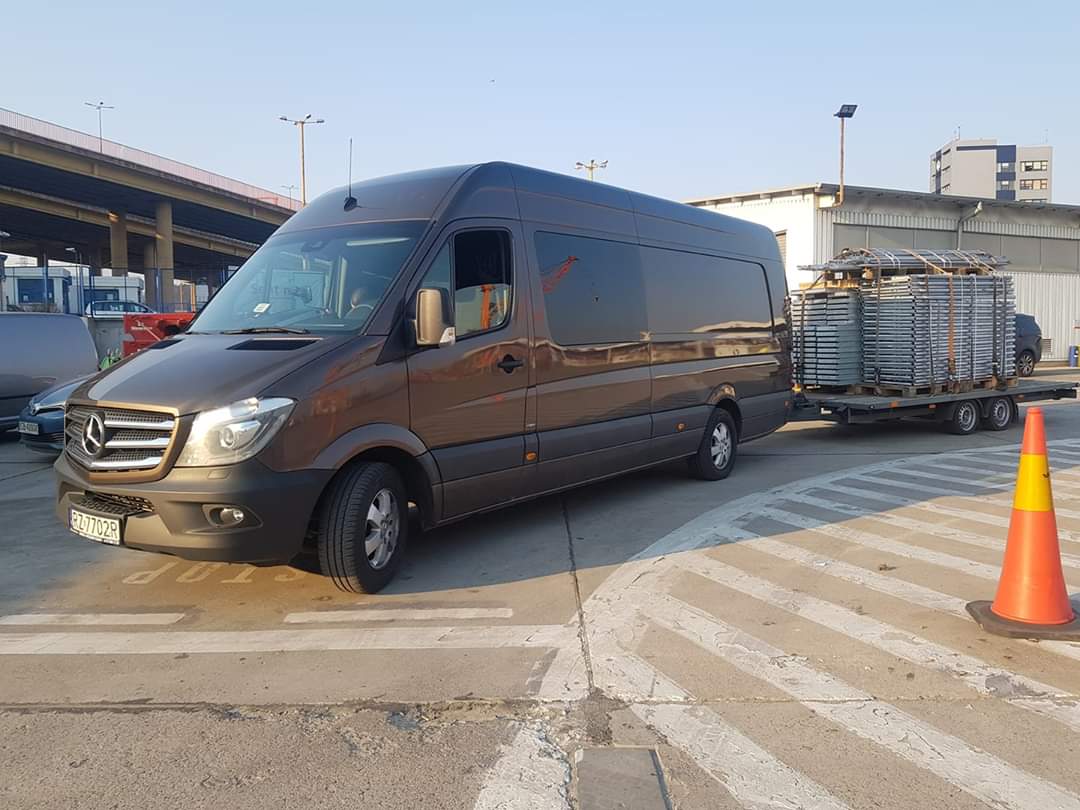 Brązowy van Mercedes-Benz z przyczepą załadowaną metalowymi konstrukcjami, zaparkowany na placu z widocznym stożkiem ostrzegawczym.