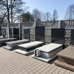 Ekspozycja różnorodnych pomników z kamienia i konglomeratu na placu z kostki brukowej, w tle betonowe ogrodzenie z dekoracyjnym wzorem cegły.