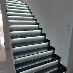 Nowoczesne schody wewnętrzne z czarnymi stopnicami i podświetleniem LED, marmurowe podstopnice, białe ściany i podłoga z połyskiem.