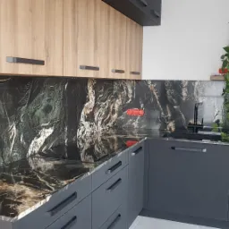 Kuchenny blat z czarnego kamienia z wyraźnym, białym i złotym użyleniem, dopasowany do ciemnoszarych szafek i drewnianych szafek wiszących, uzupełniony o rośliny doniczkowe na półce.