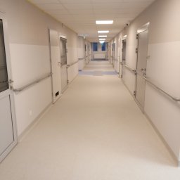 FLOOR-POL - Długi korytarz w budynku użyteczności publicznej z jasną, jednolitą posadzką, białymi ścianami z poziomym pasem w kolorze beżowym i aluminiowymi poręczami, oraz rzędem identycznych drzwi po obu...