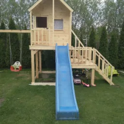 Drewniany domek dla dzieci na podwyższeniu z niebieską zjeżdżalnią i huśtawką na tle zielonego trawnika i wysokich drzew.