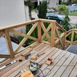 FLOOR-POL - Drewniany taras z balustradą w trakcie budowy, widoczne narzędzia pracy: młotek, poziomica, skrzynka z gwoździami, przedłużacz i transformator, w tle zabudowa jednorodzinna i zieleń.