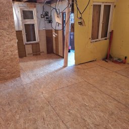 FLOOR-POL - Wnętrze w trakcie remontu z ułożoną podłogą z płyt OSB, widoczne okna, żółta ściana, wystające kable i narzędzia w tle.