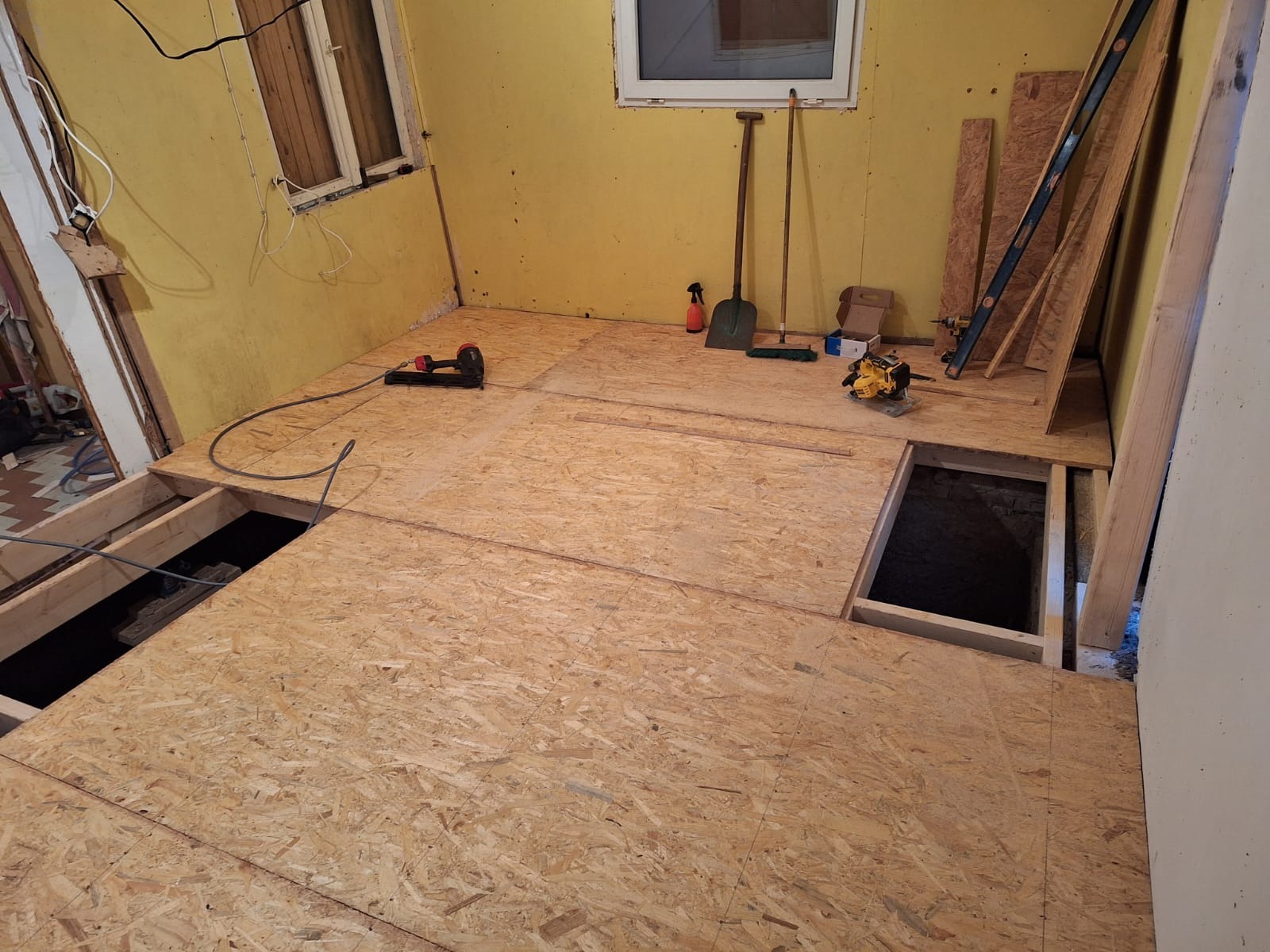 Montaż podłogi z płyt OSB w pomieszczeniu z żółtymi ścianami, widoczne otwory w podłodze, narzędzia (zszywacz, piła), poziomica, deski i akcesoria malarskie.