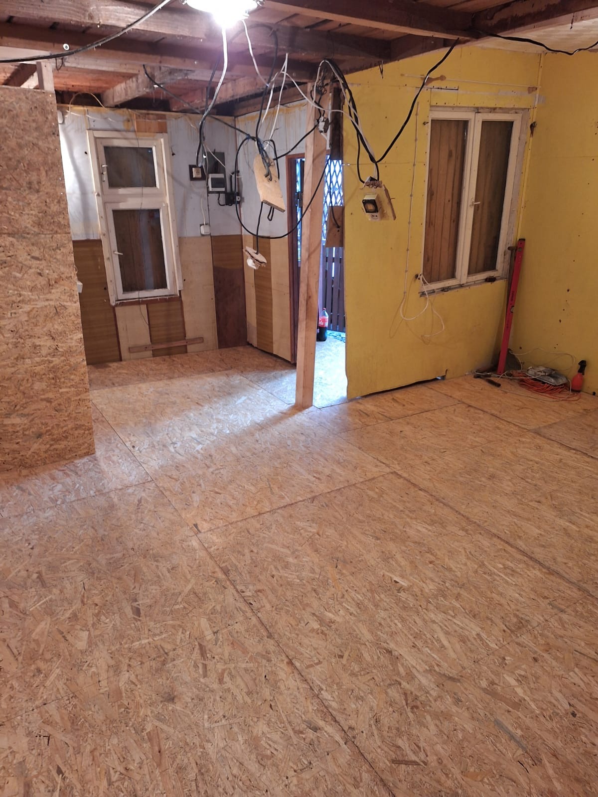 Wnętrze w trakcie remontu z ułożoną podłogą z płyt OSB, widoczne okna, żółta ściana, wystające kable i narzędzia w tle.
