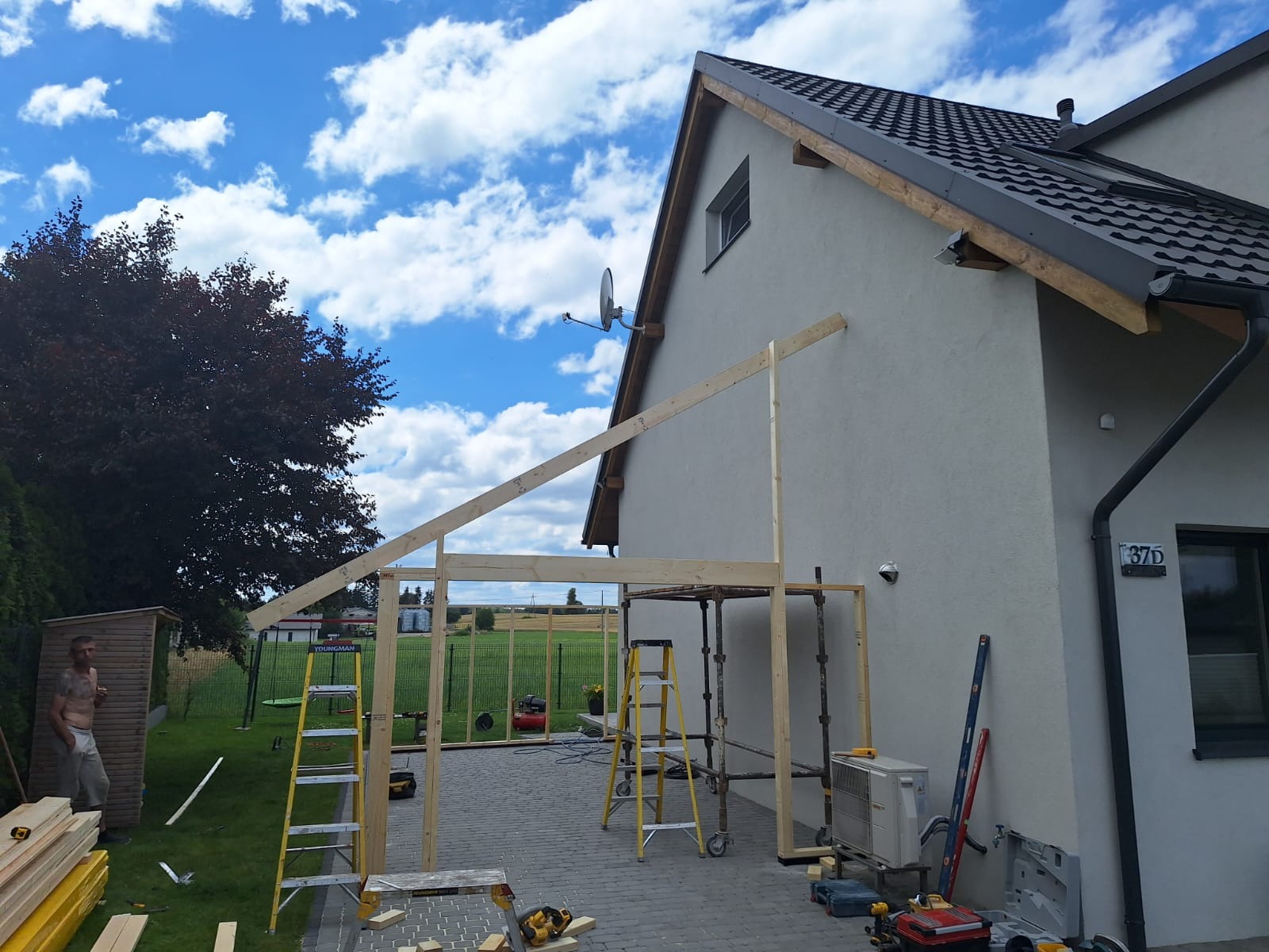 Budowa drewnianej konstrukcji przylegającej do domu jednorodzinnego z szarą elewacją, widoczne rusztowania, drabiny, narzędzia i materiały budowlane na brukowanym podłożu, w tle zielony trawnik...