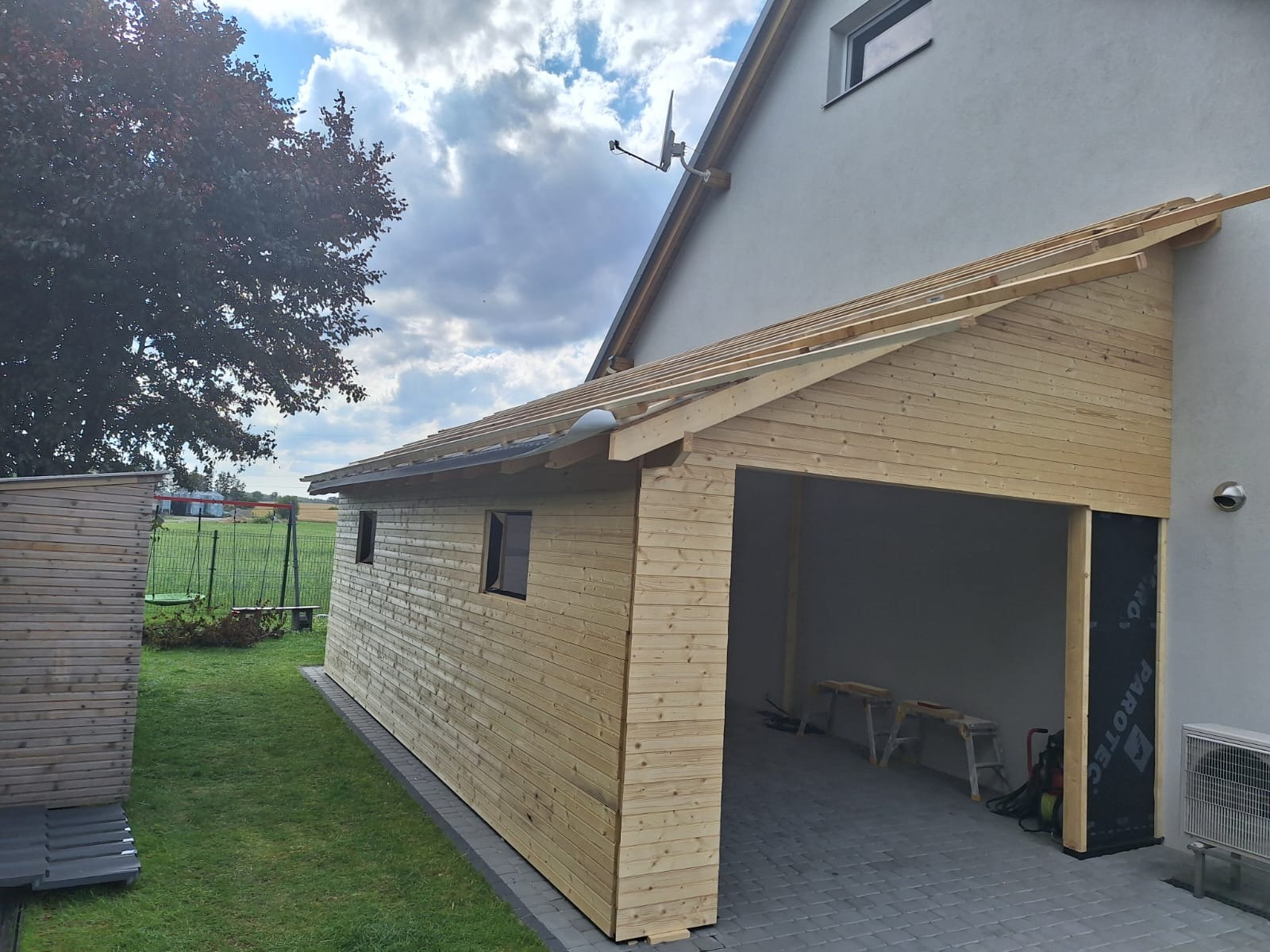 Budowa drewnianej konstrukcji garażu z elementami domu szkieletowego, widoczne krokwie dachu i deski elewacyjne, obok mała drewniana szopa na narzędzia.