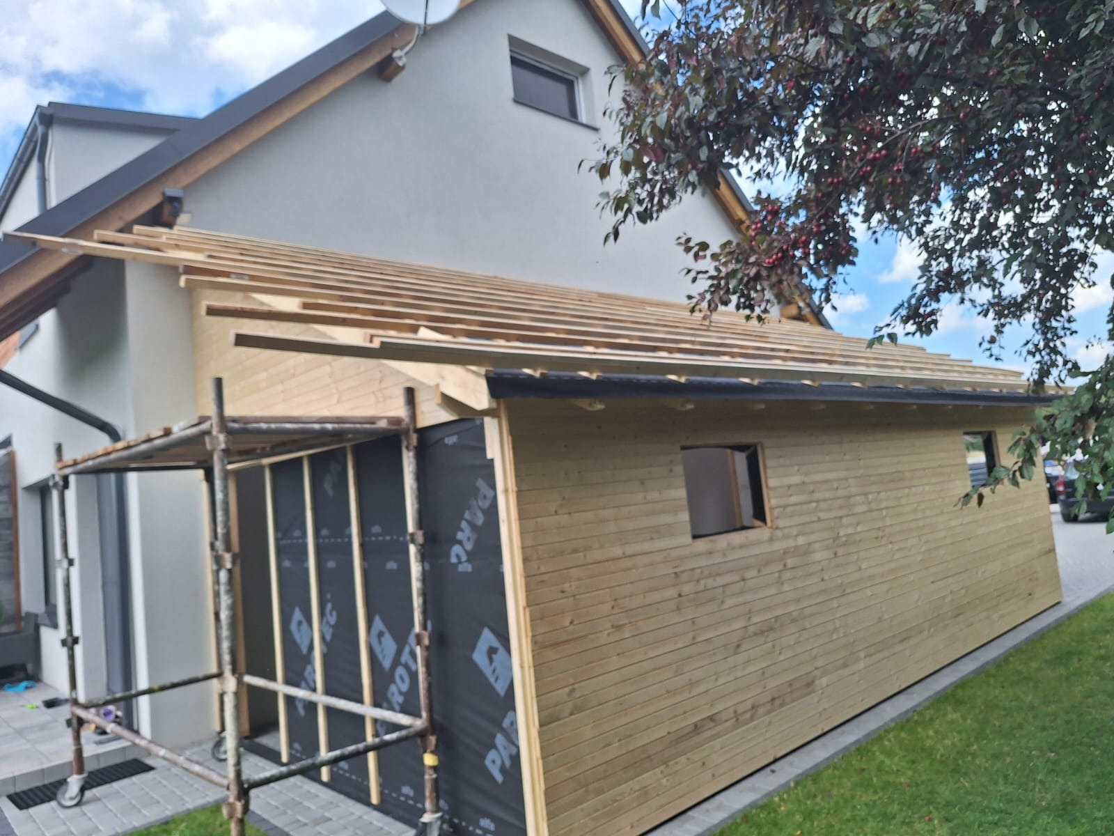 Budowa domu szkieletowego w Starogardzie Gdańskim: widok z zewnątrz na konstrukcję z drewna, częściowo obłożoną folią paroizolacyjną, z widocznym rusztowaniem i przygotowywanym dachem.