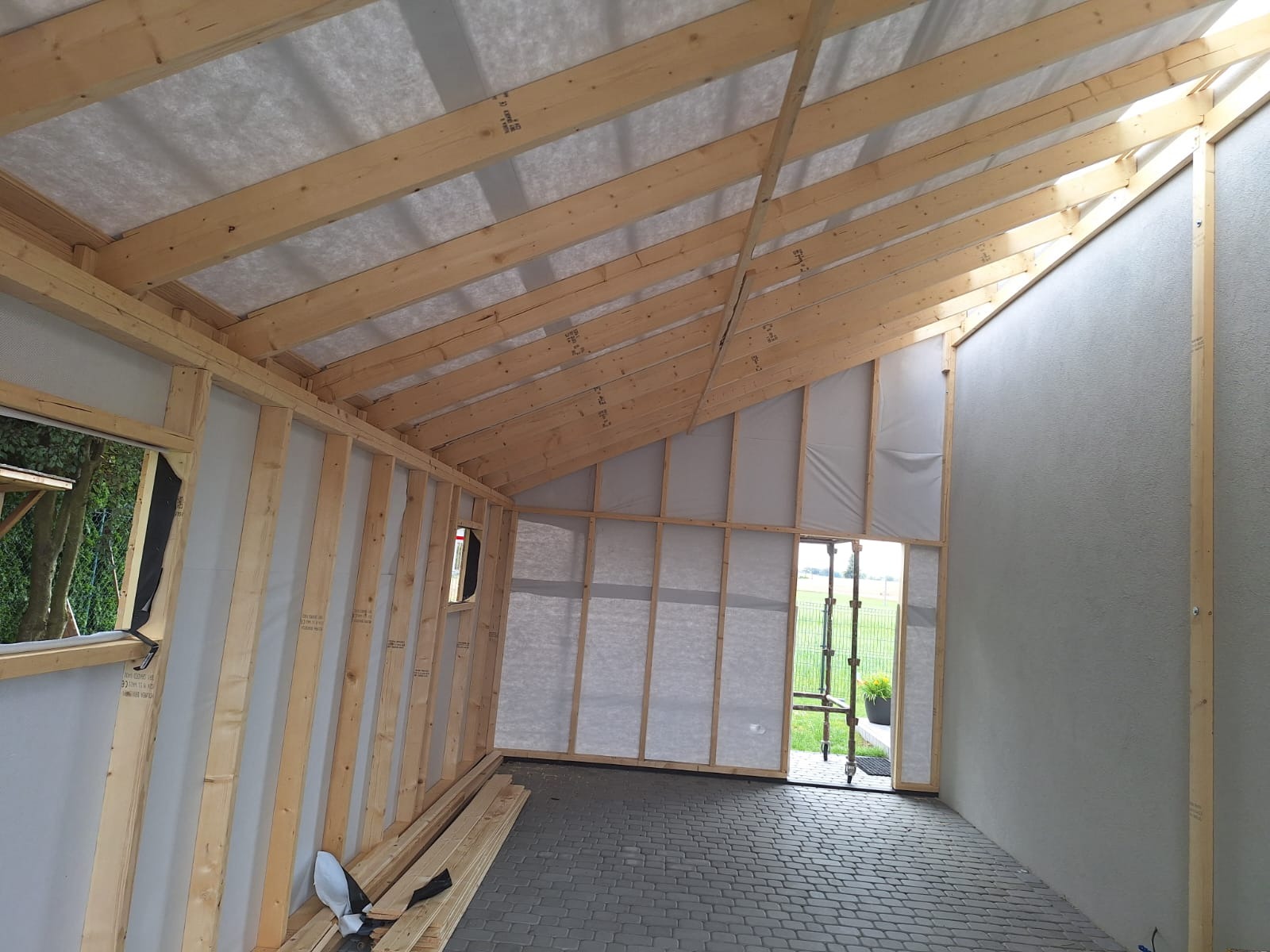 Wnętrze domu szkieletowego w trakcie budowy: widoczne drewniane ramy ścian, konstrukcja dachu z izolacją, szara ściana zewnętrzna, podłoga z szarej kostki brukowej oraz widok na zielone otoczenie...