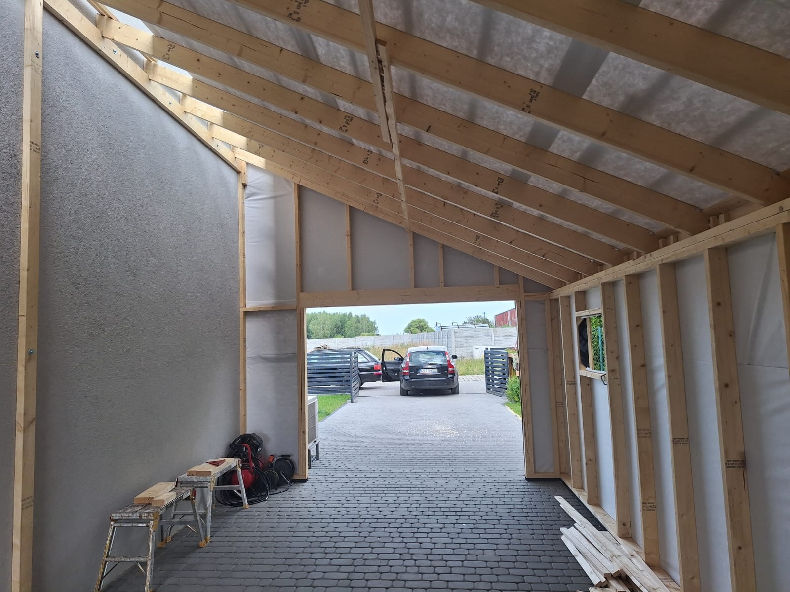 Wnętrze w trakcie budowy domu szkieletowego: widoczne drewniane ramy konstrukcyjne, ocieplenie ścian, szara ściana z gładzią, widok na podjazd z kostki brukowej i zaparkowane samochody.