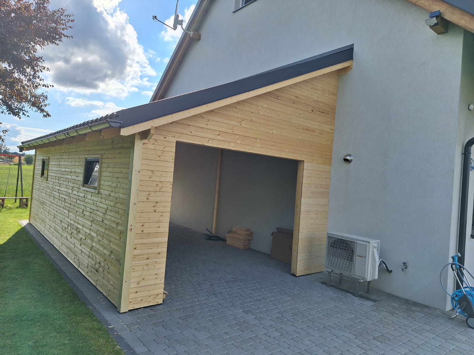 Nowo wybudowana, drewniana wiata garażowa przylegająca do domu, z widocznym fragmentem elewacji i klimatyzatora zewnętrznego.