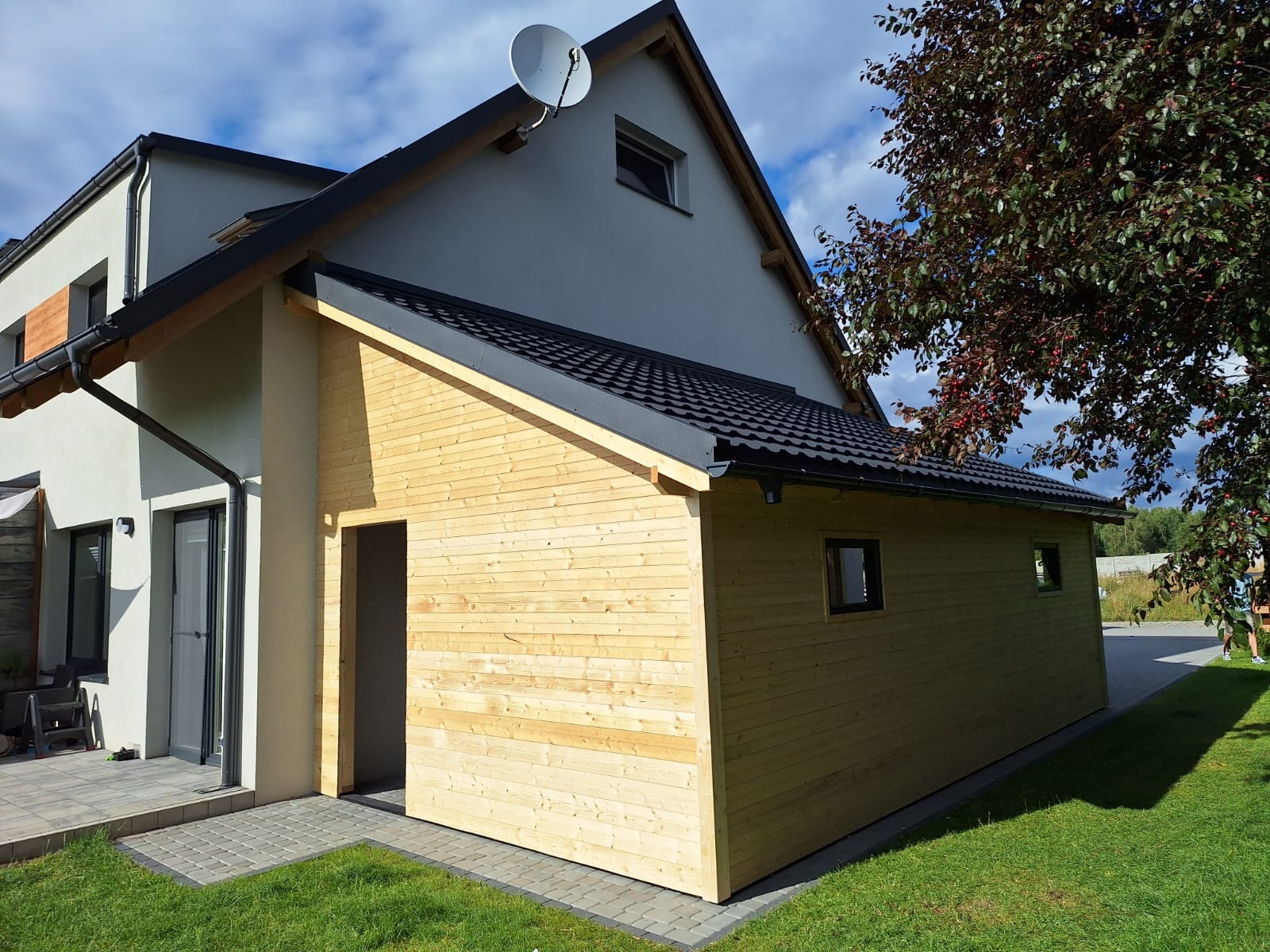 Nowo wybudowany, drewniany budynek gospodarczy przylegający do domu jednorodzinnego z szarą elewacją, czarnym dachem i satelitą na dachu, otoczony trawnikiem i drzewem.