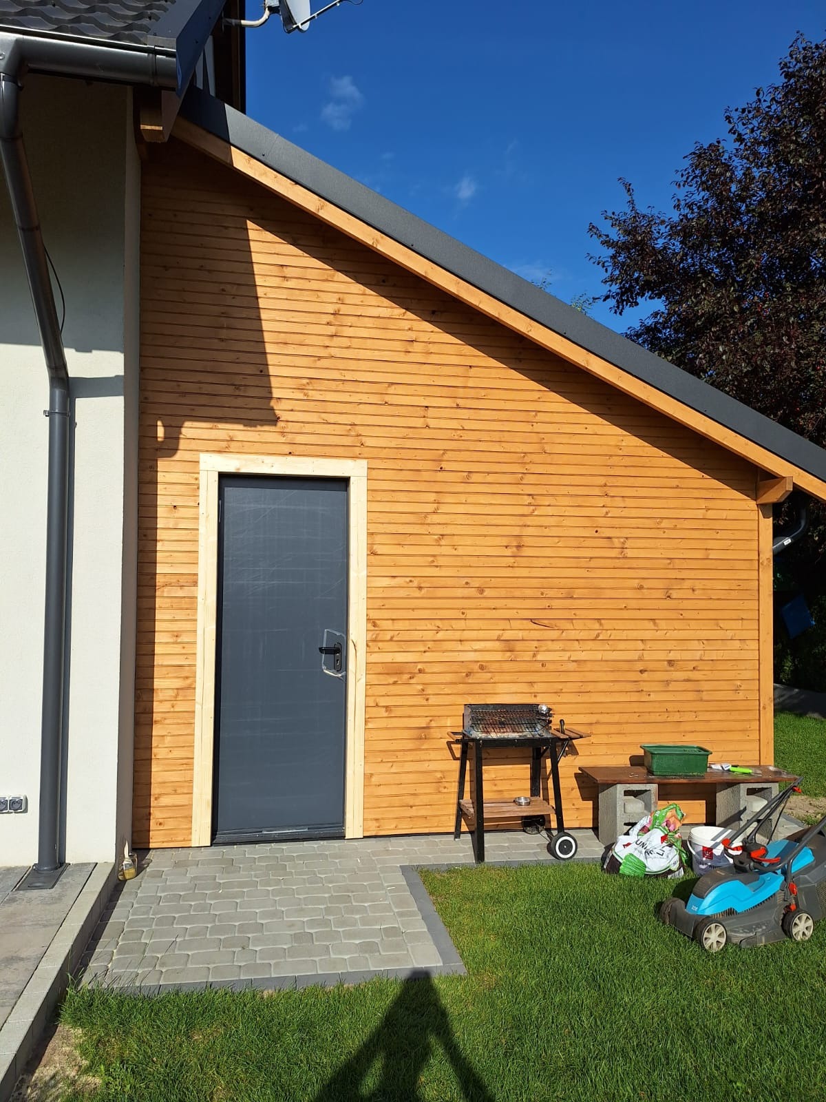 Drewniana konstrukcja budynku z szarą, metalową stolarką drzwiową, przylegająca do murowanej części domu, z grillem i kosiarką na trawniku.