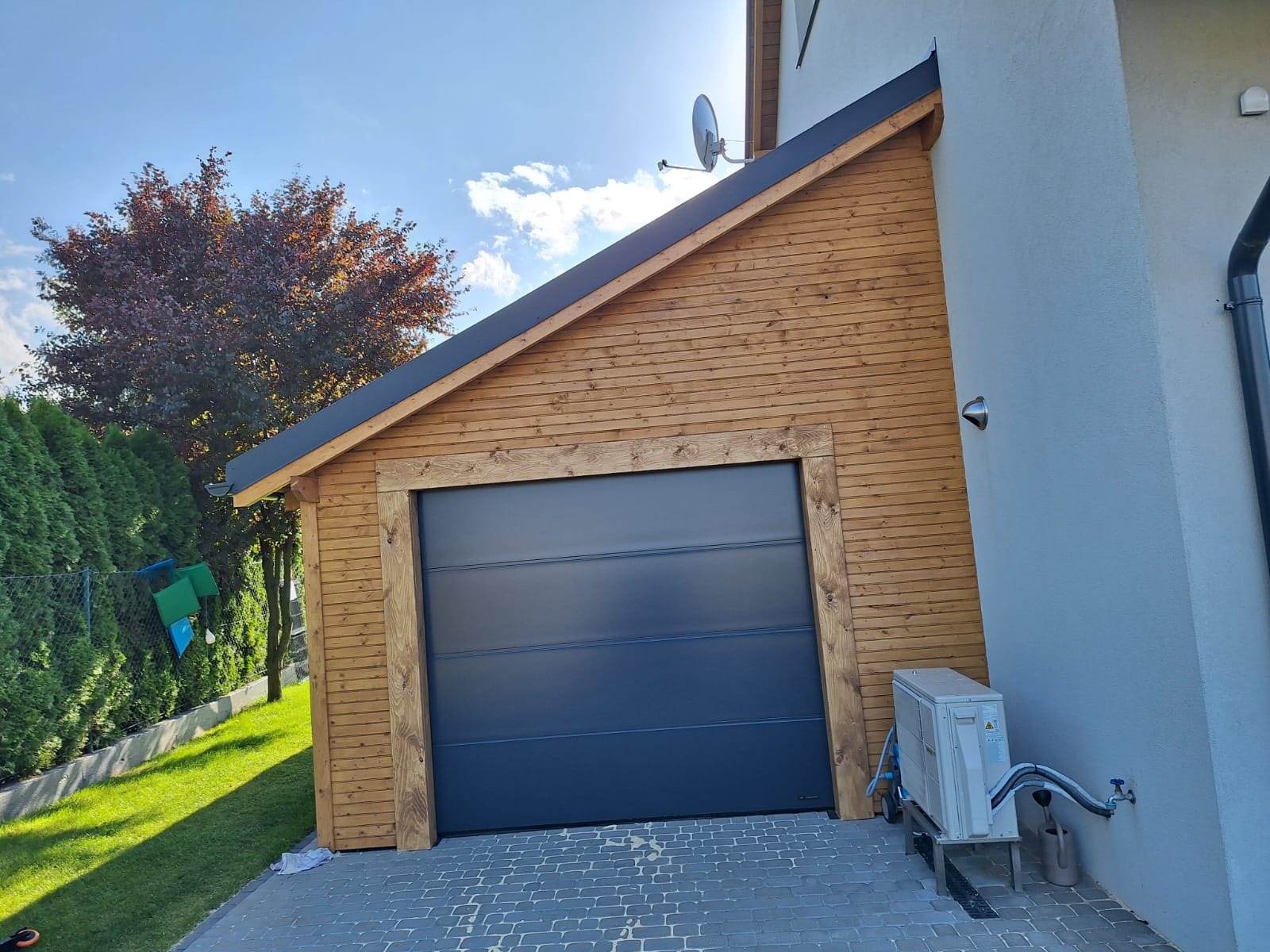 Nowoczesny, drewniany garaż z segmentową bramą w kolorze antracytowym, przylegający do jasnego budynku mieszkalnego, widoczny klimatyzator na zewnątrz.