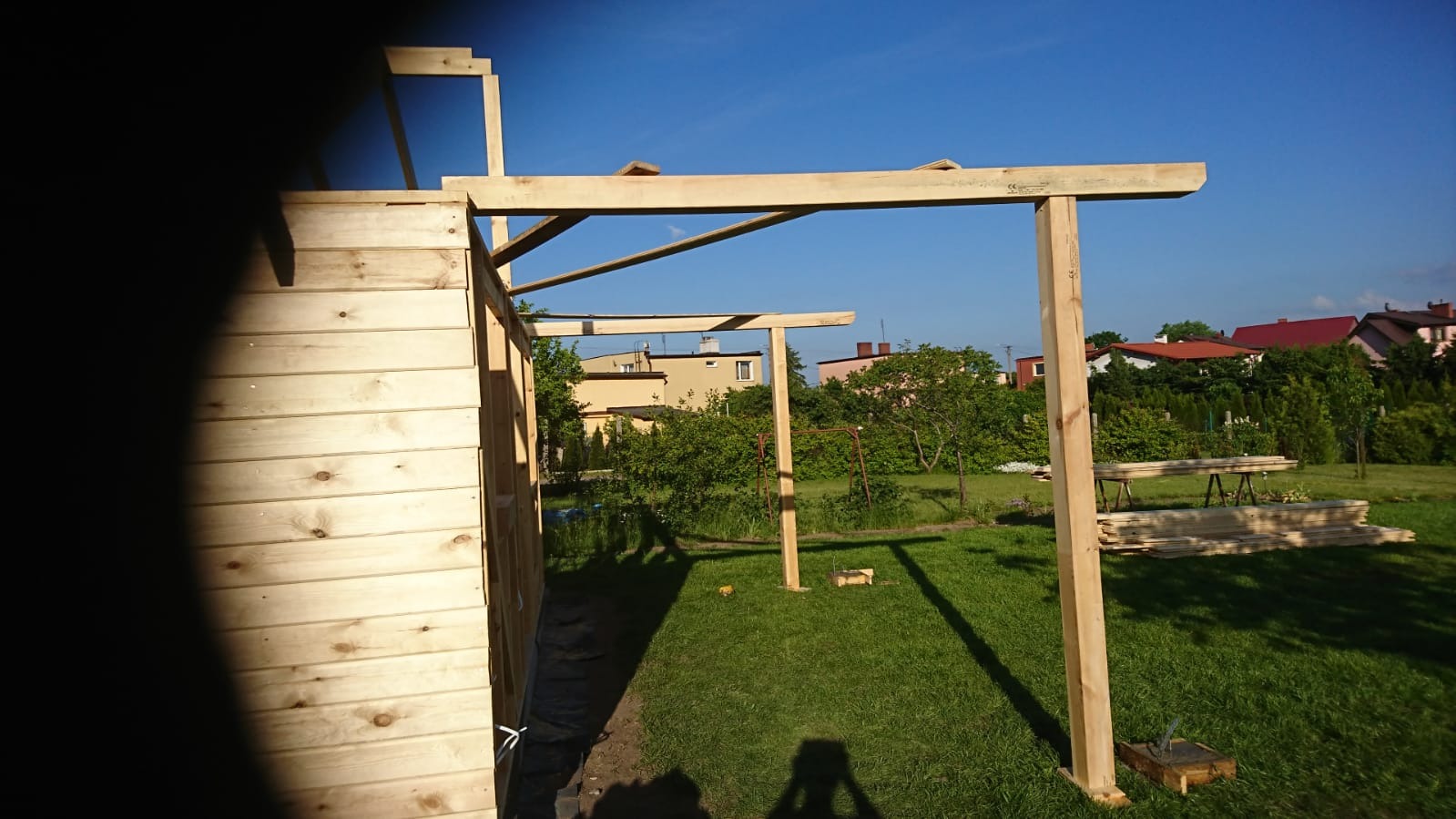 Wznoszenie konstrukcji szkieletowej z surowego drewna na zielonym trawniku w słoneczny dzień, widoczne deski przygotowane do montażu.