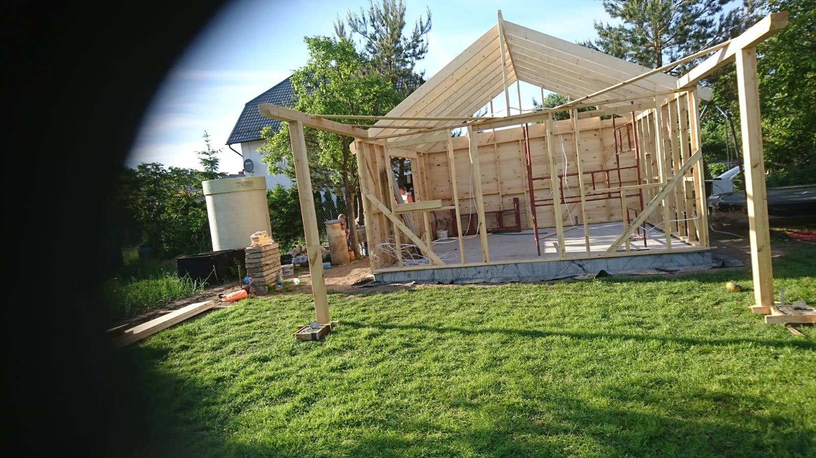 Wznoszenie szkieletu drewnianego domu na trawniku, widoczne elementy konstrukcyjne ścian i dachu, w tle budynek mieszkalny i drzewa.
