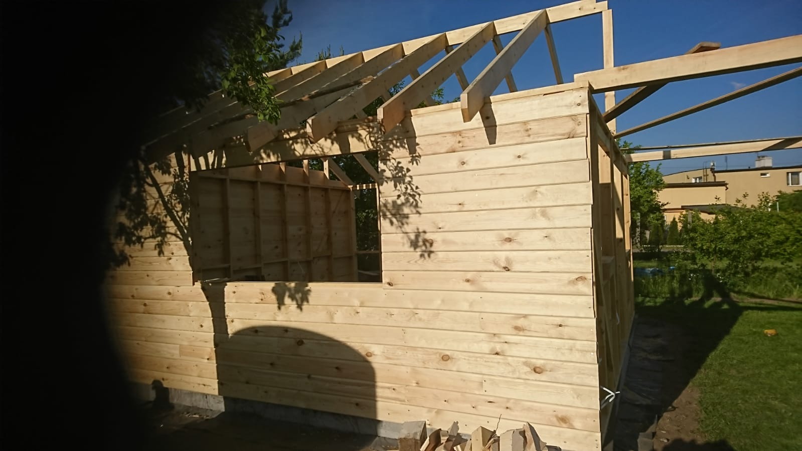 Wznoszenie szkieletowej konstrukcji domu z widocznymi drewnianymi belkami stropowymi i ścianami obitymi deskami, na tle zieleni ogrodu i błękitnego nieba.