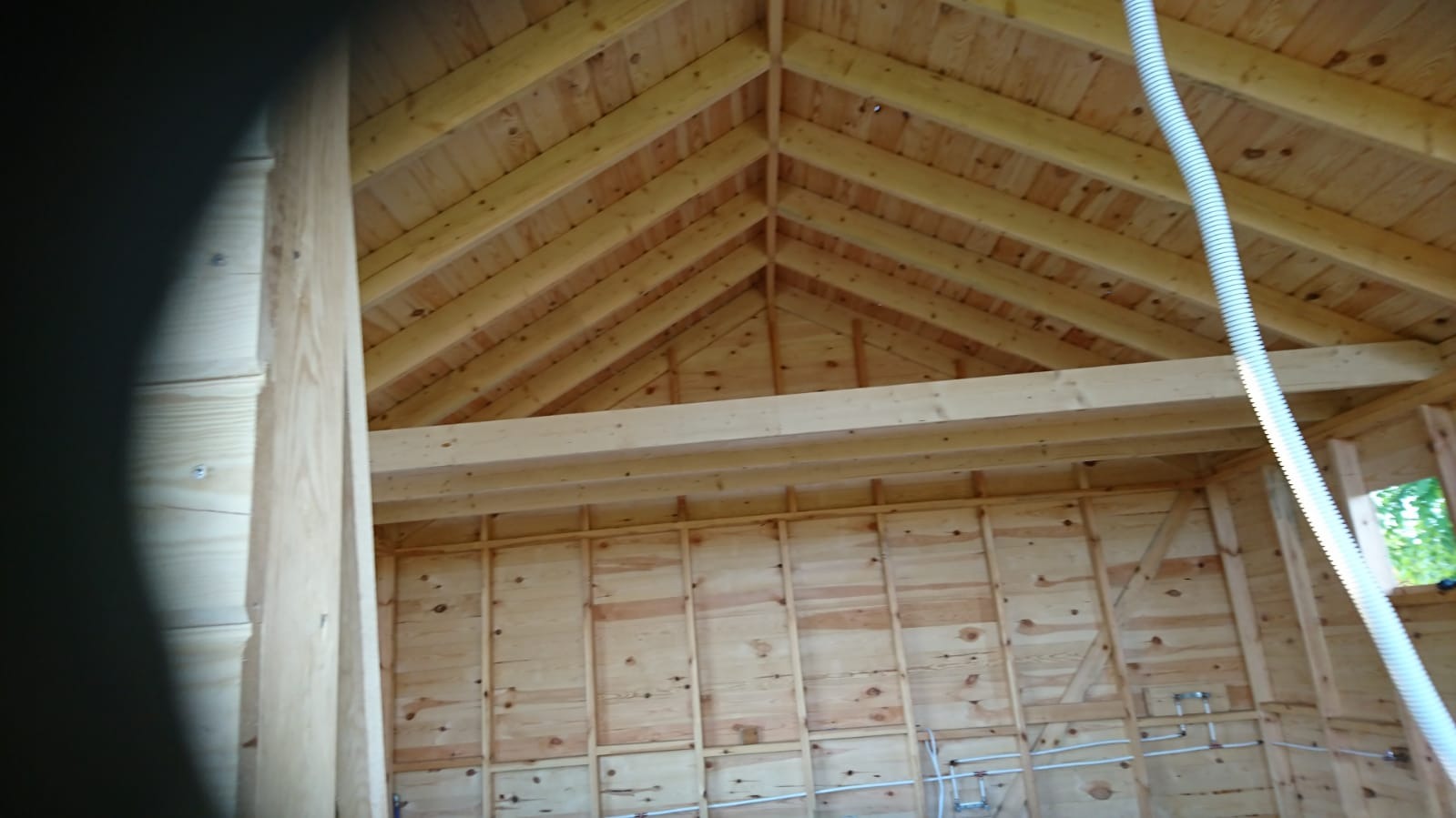 Wnętrze domu szkieletowego w trakcie budowy, widoczne drewniane elementy konstrukcyjne ścian i dachu, biała rura wentylacyjna zwisająca z sufitu, instalacja wodna na ścianie.