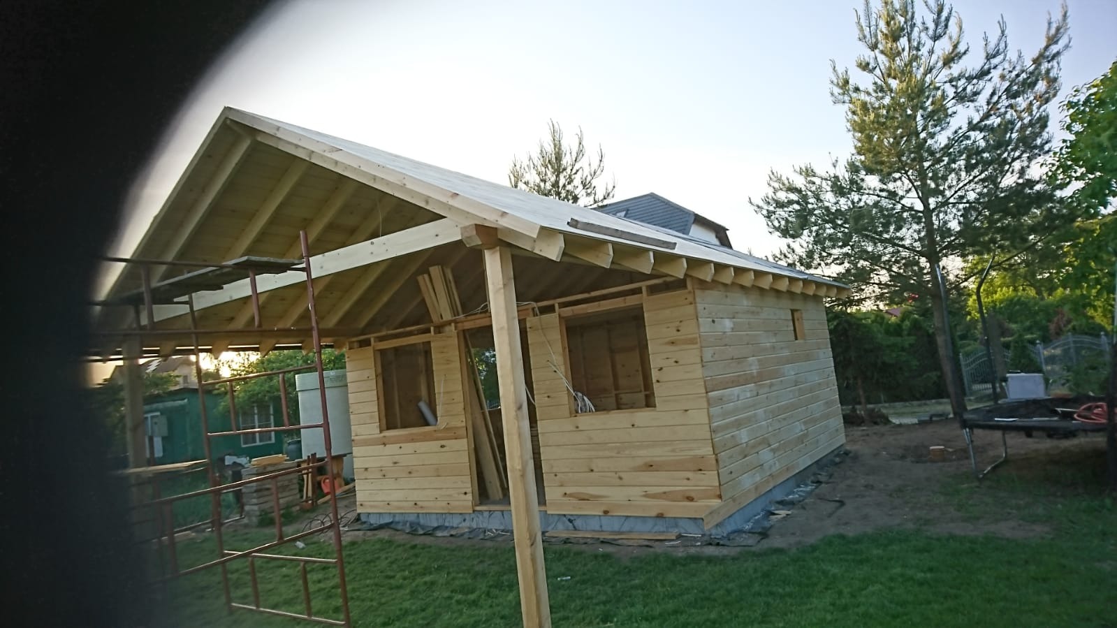 Wznoszona konstrukcja drewniana domu szkieletowego, widoczne ściany z desek, więźba dachowa i rusztowanie, w tle zieleń ogrodu.