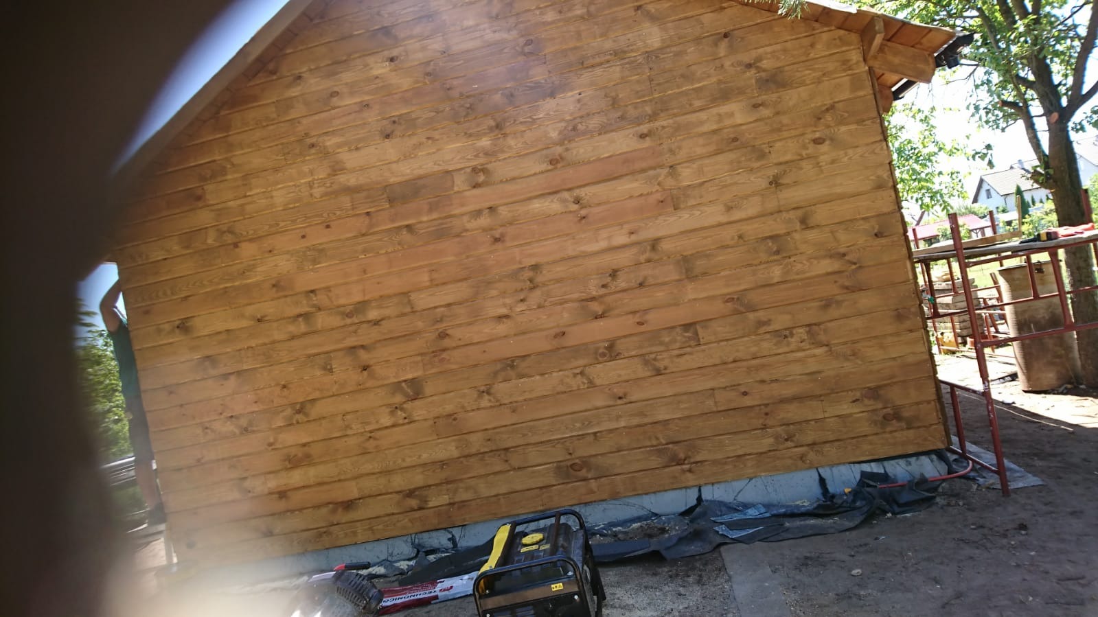 Świeżo pomalowana, drewniana ściana domu szkieletowego z widocznym agregatem prądotwórczym i rusztowaniem w tle.