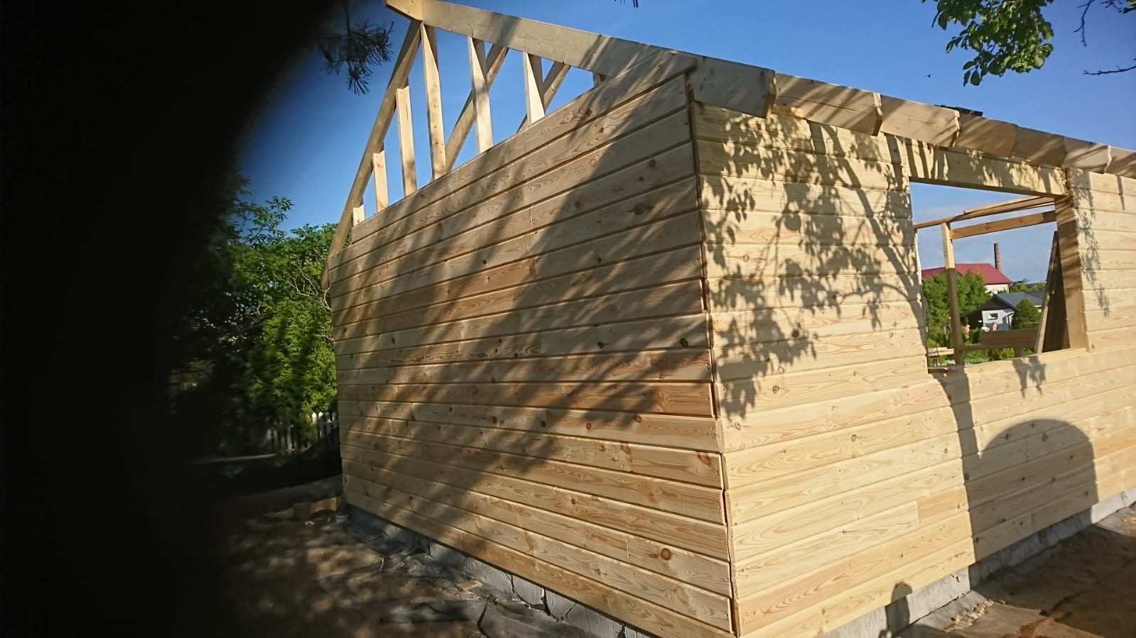 Wznoszenie surowej konstrukcji domu z drewnianych bali na fundamencie, widoczne elementy więźby dachowej i otwory okienne, cień drzewa na elewacji.