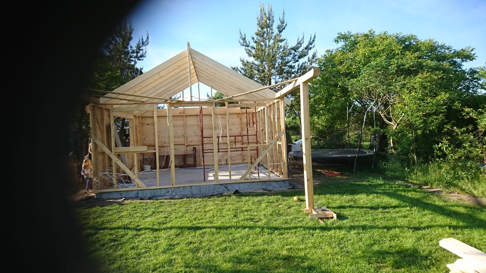 Konstrukcja szkieletowa domu w trakcie budowy, widoczne drewniane elementy ścian, dachu i stropu, na zewnątrz trawnik i drzewa, w tle trampolina