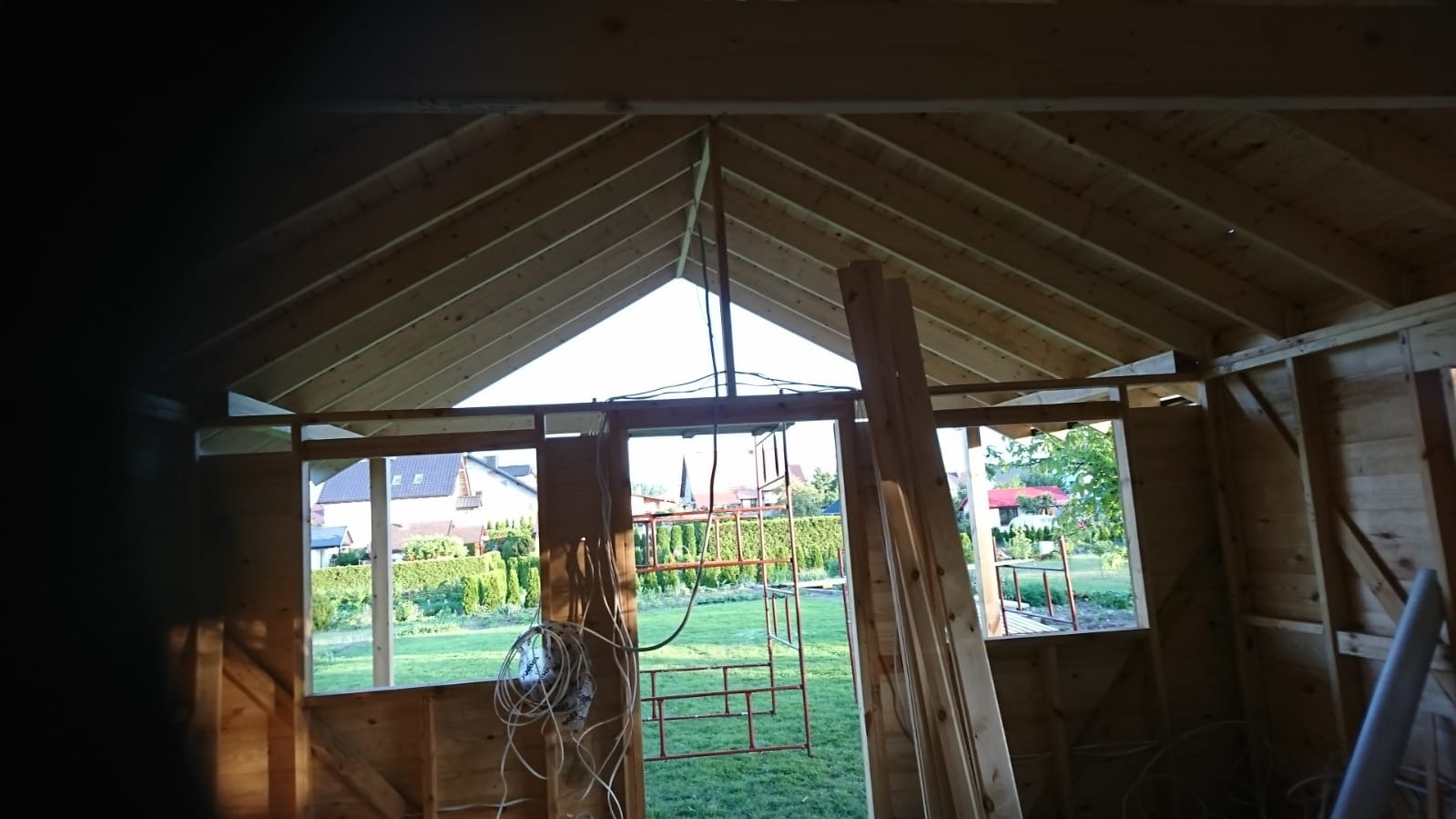 Wnętrze surowego drewnianego domku szkieletowego w trakcie budowy, widok na ogród przez otwory okienne i drzwiowe, widoczne elementy konstrukcyjne dachu i ścian.