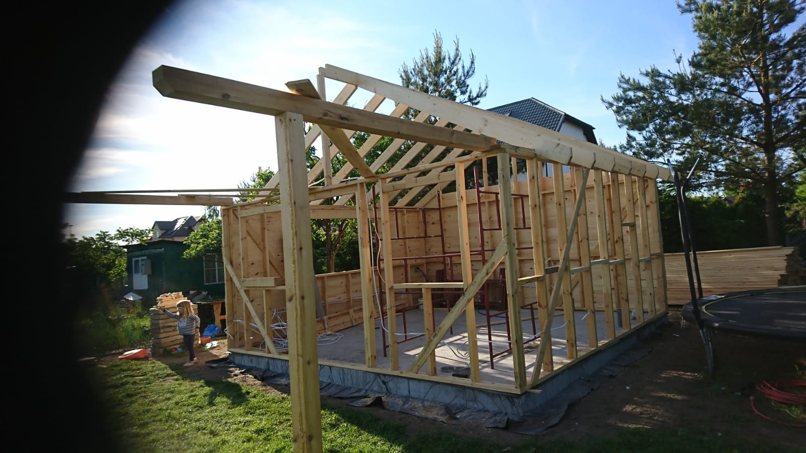 Wznoszenie konstrukcji szkieletowej domu z widocznymi elementami drewnianymi, izolacją fundamentów i częściowo ułożonymi deskami na podłogę, z dziewczynką w tle i trampoliną w ogrodzie.