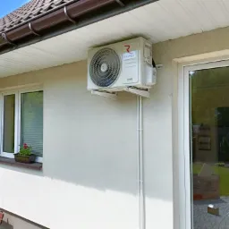 Zewnętrzna jednostka klimatyzacji Rotenso Ukura zamontowana na jasnej ścianie domu, widoczne rury prowadzące w d&oacute;ł, okno z kwiatami i odbicie ogrodu w szybie drzwi balkonowych.